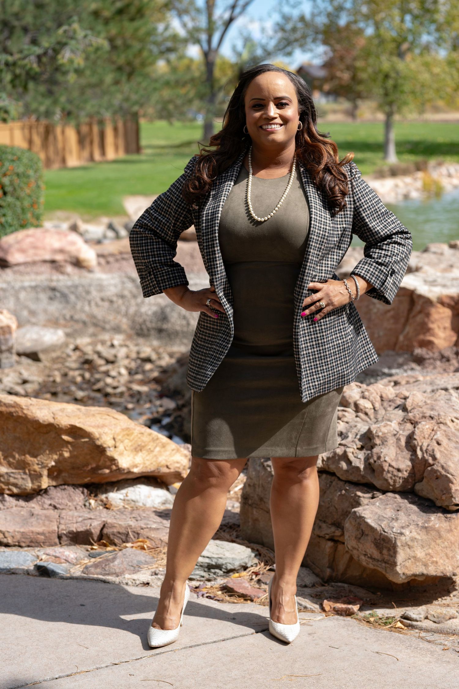 Professional woman in houndstooth blazer and olive dress poses confidently outdoors near rocky water feature.