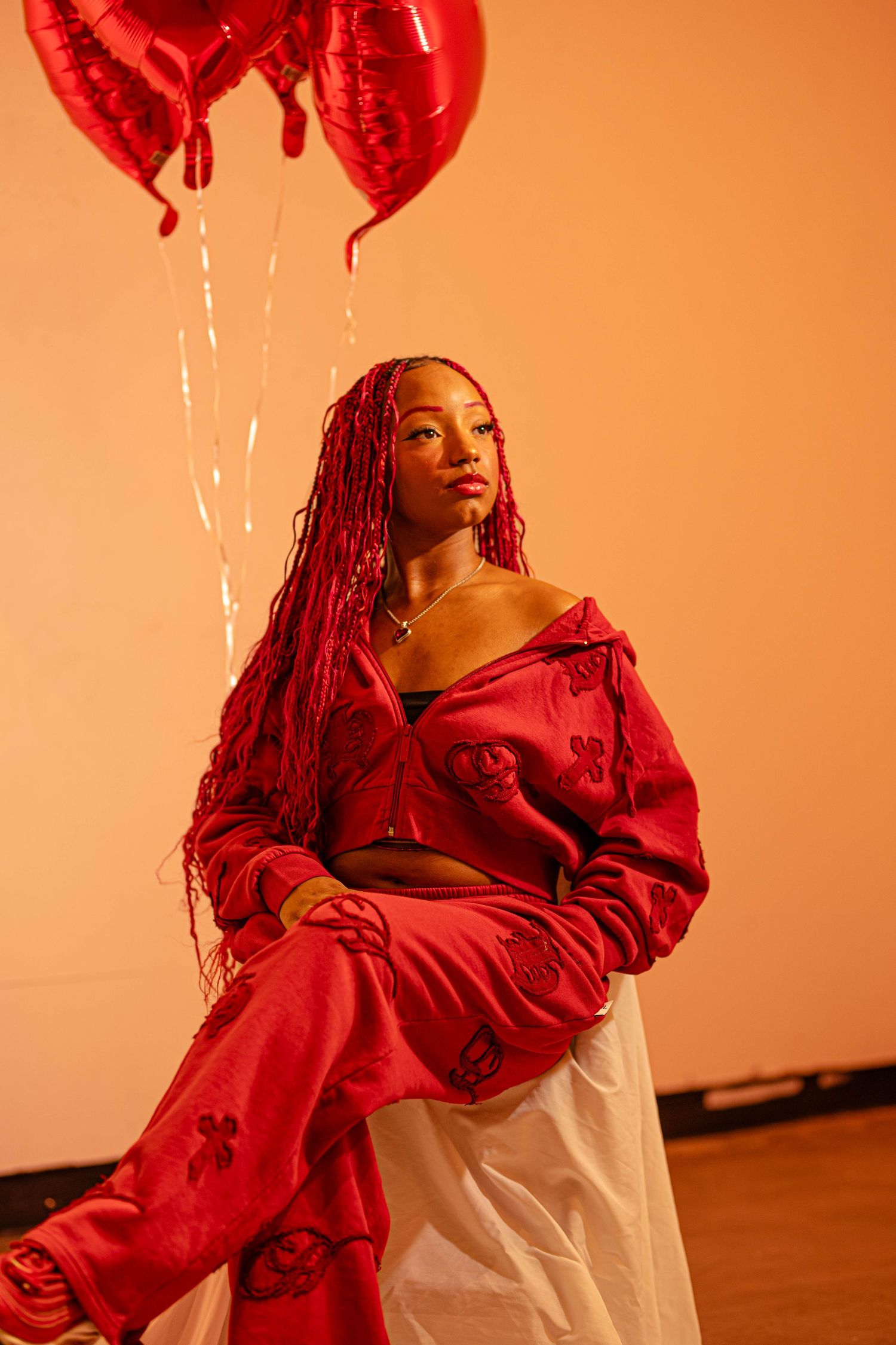 Woman in red outfit with red braids seated under heart-shaped balloons in a studio portrait with soft lighting.