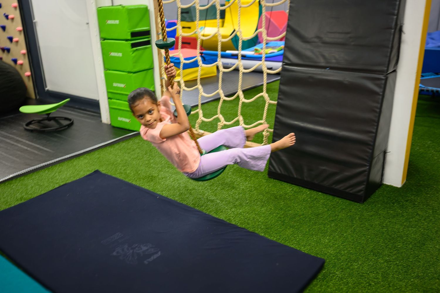 Child swinging on rope net in bright indoor pediatric therapy and play gym.