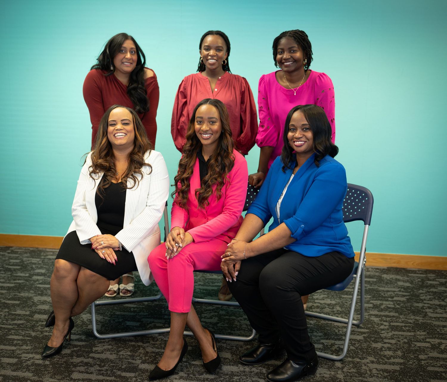 Six professional women pose together smiling for a group photo in a bright office environment with teal walls.