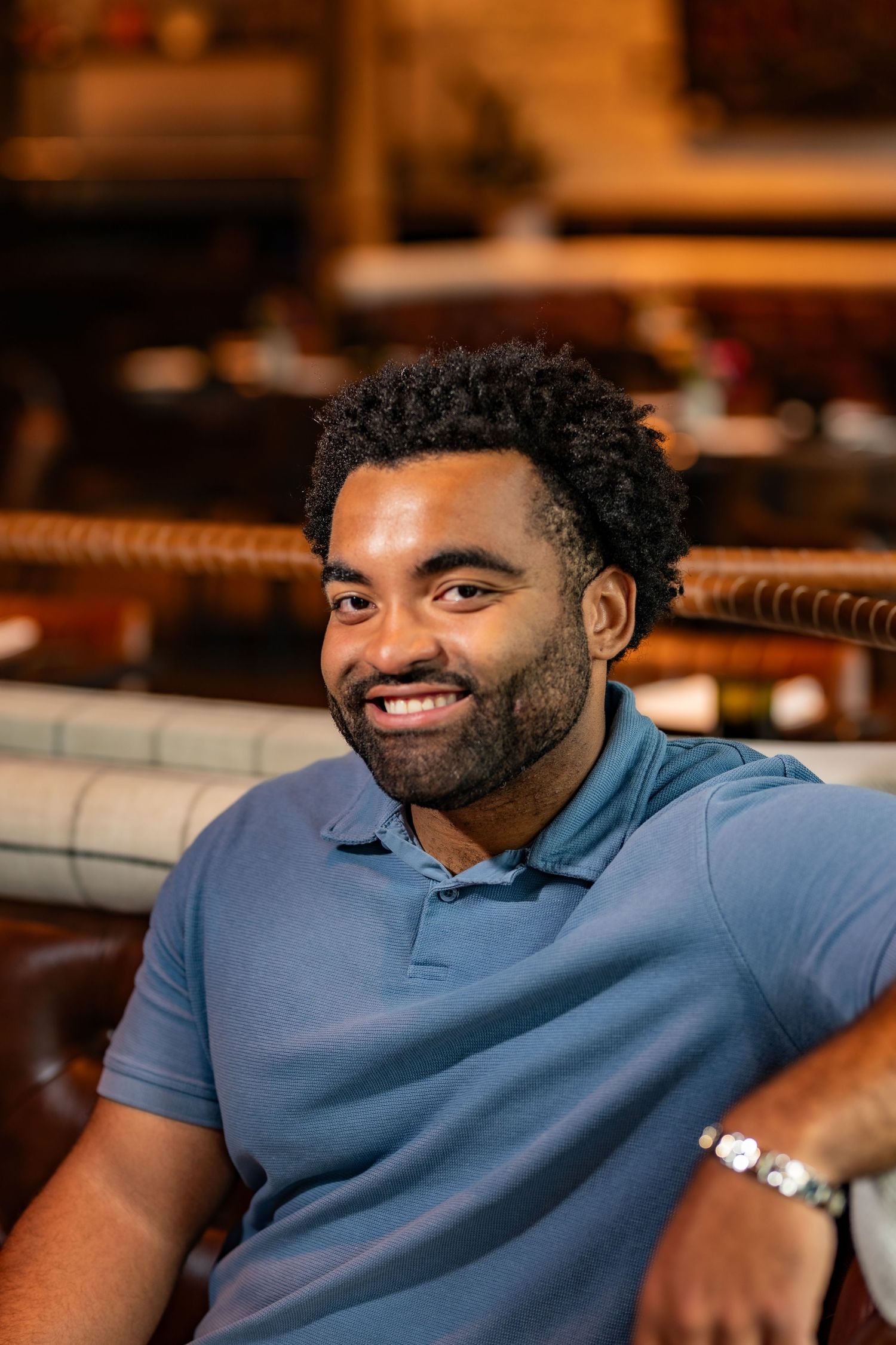 Smiling young Black man with beard wearing blue polo shirt relaxing in warm-toned restaurant.