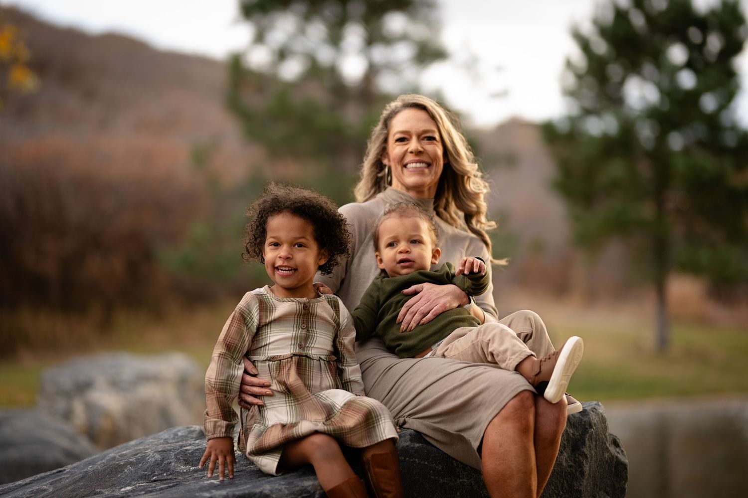 A smiling mother sits on a rock outdoors with her two young children in autumn.