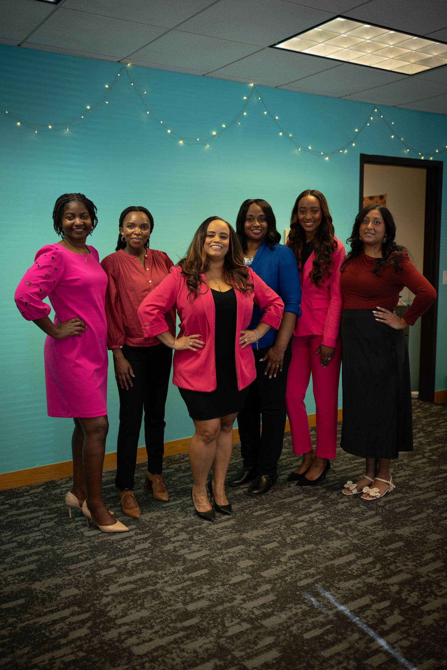 Six women in vibrant outfits pose confidently against a teal wall with string lights.