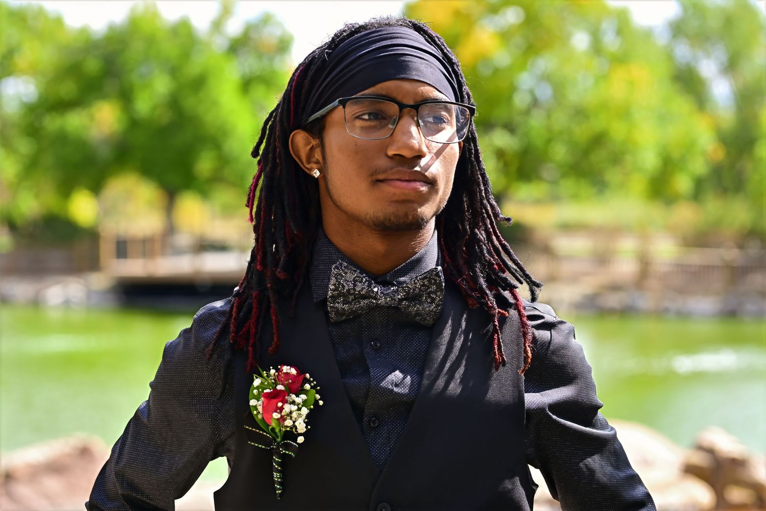 Young man with red-tipped dreadlocks wearing black suit and floral boutonniere at outdoor prom event.