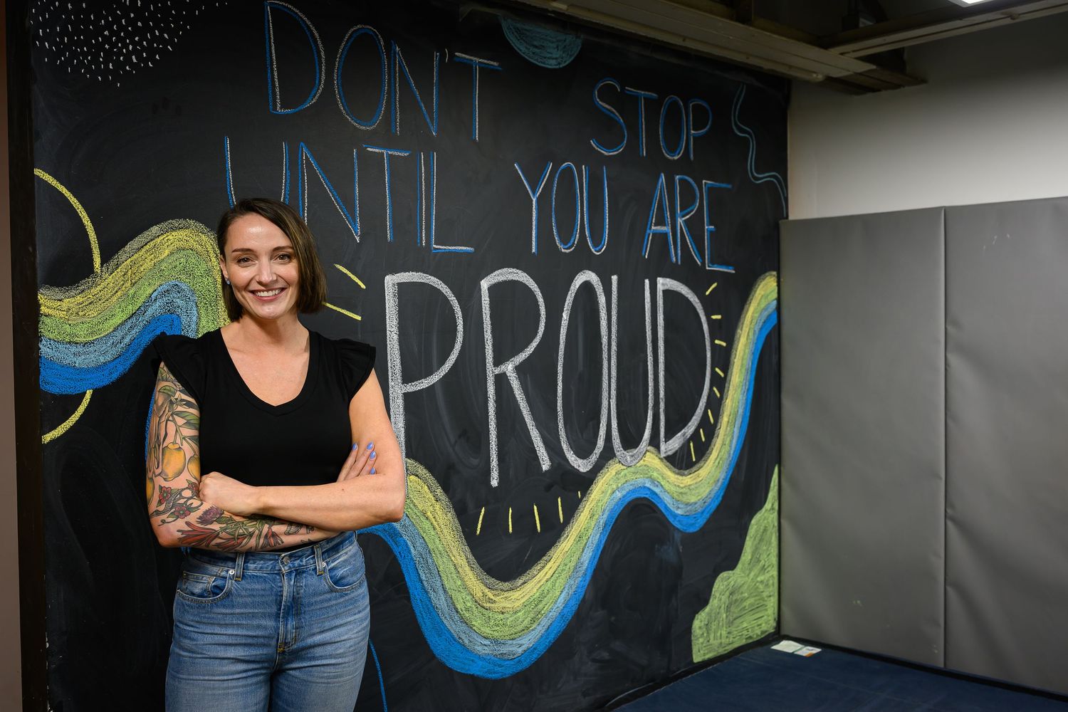 Woman with tattoos posing before motivational chalkboard sign in gym.