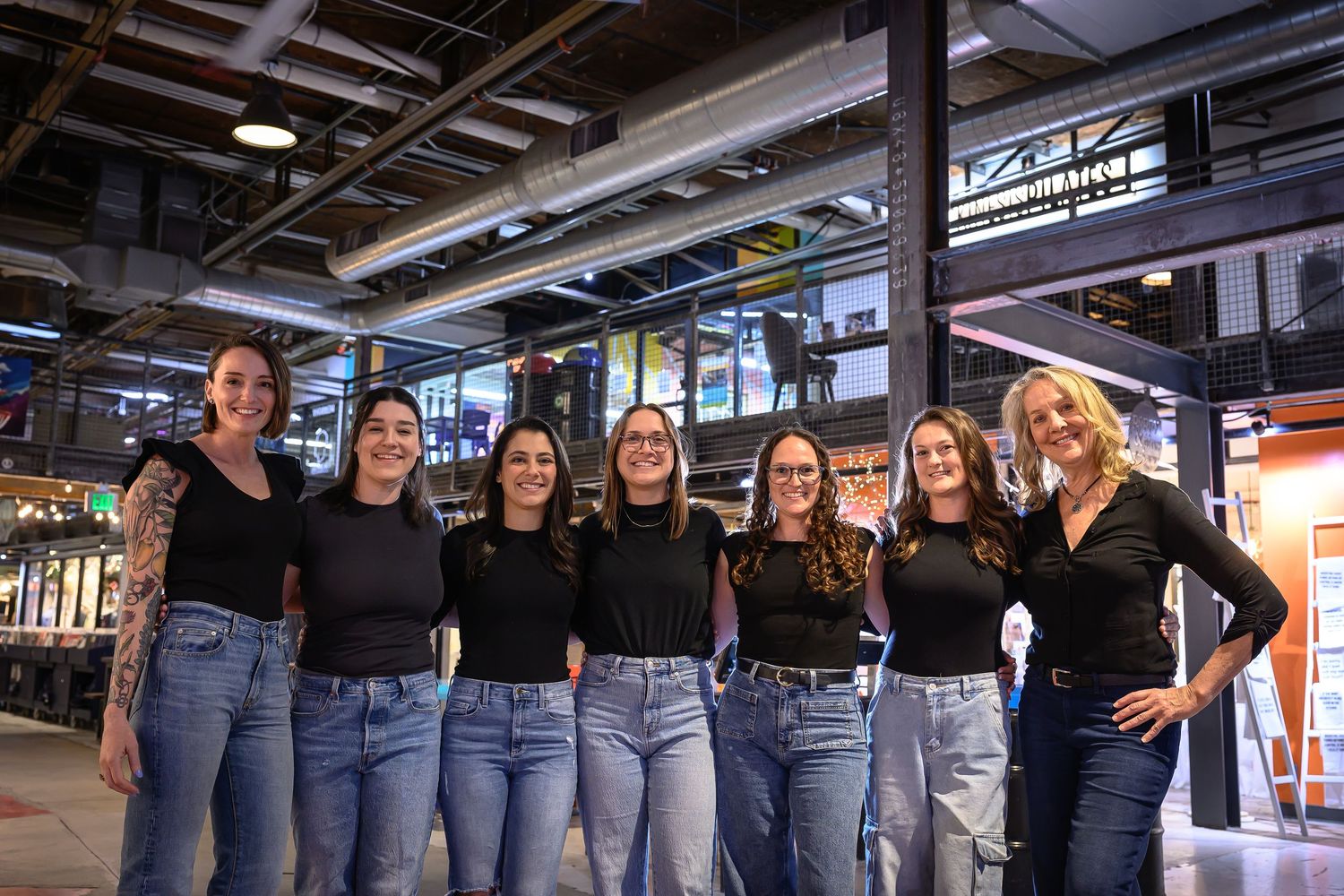Seven women in black tops and jeans smiling together in a row inside an industrial venue.