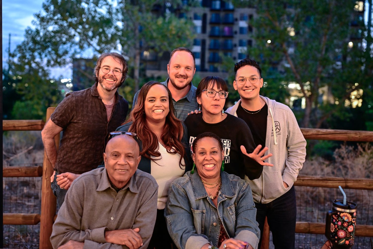 A group of seven people pose together outdoors near a wooden fence at twilight in an urban setting.