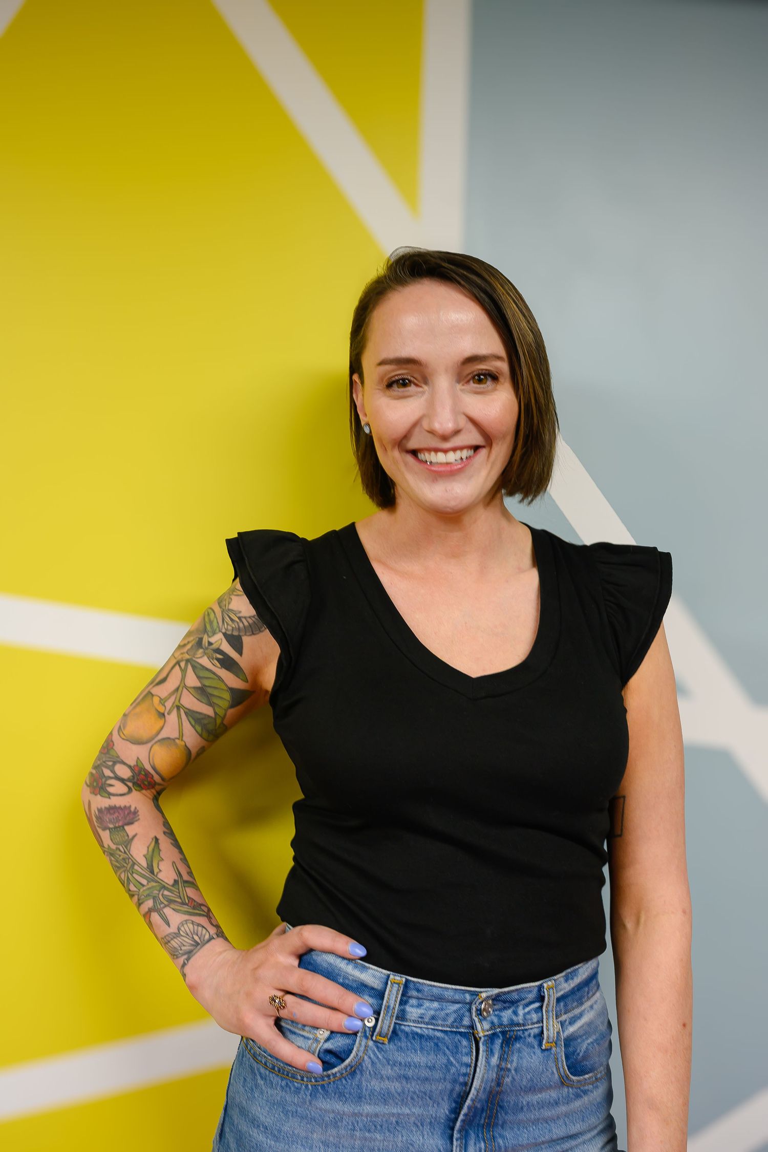 Smiling woman with floral sleeve tattoo in black ruffled top and jeans standing against yellow geometric wall.