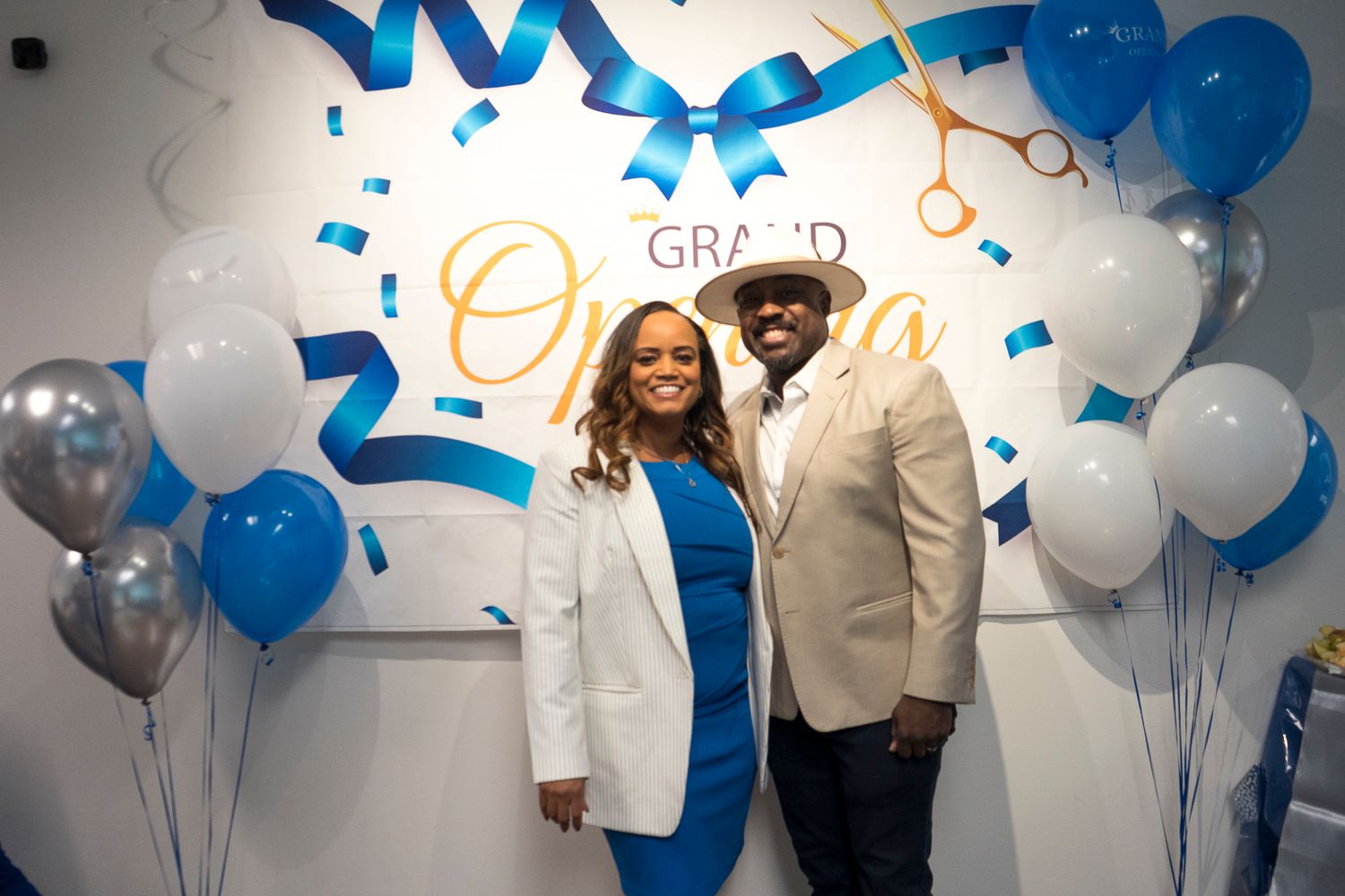 A smiling couple poses together in front of a Grand Opening sign with blue balloons.