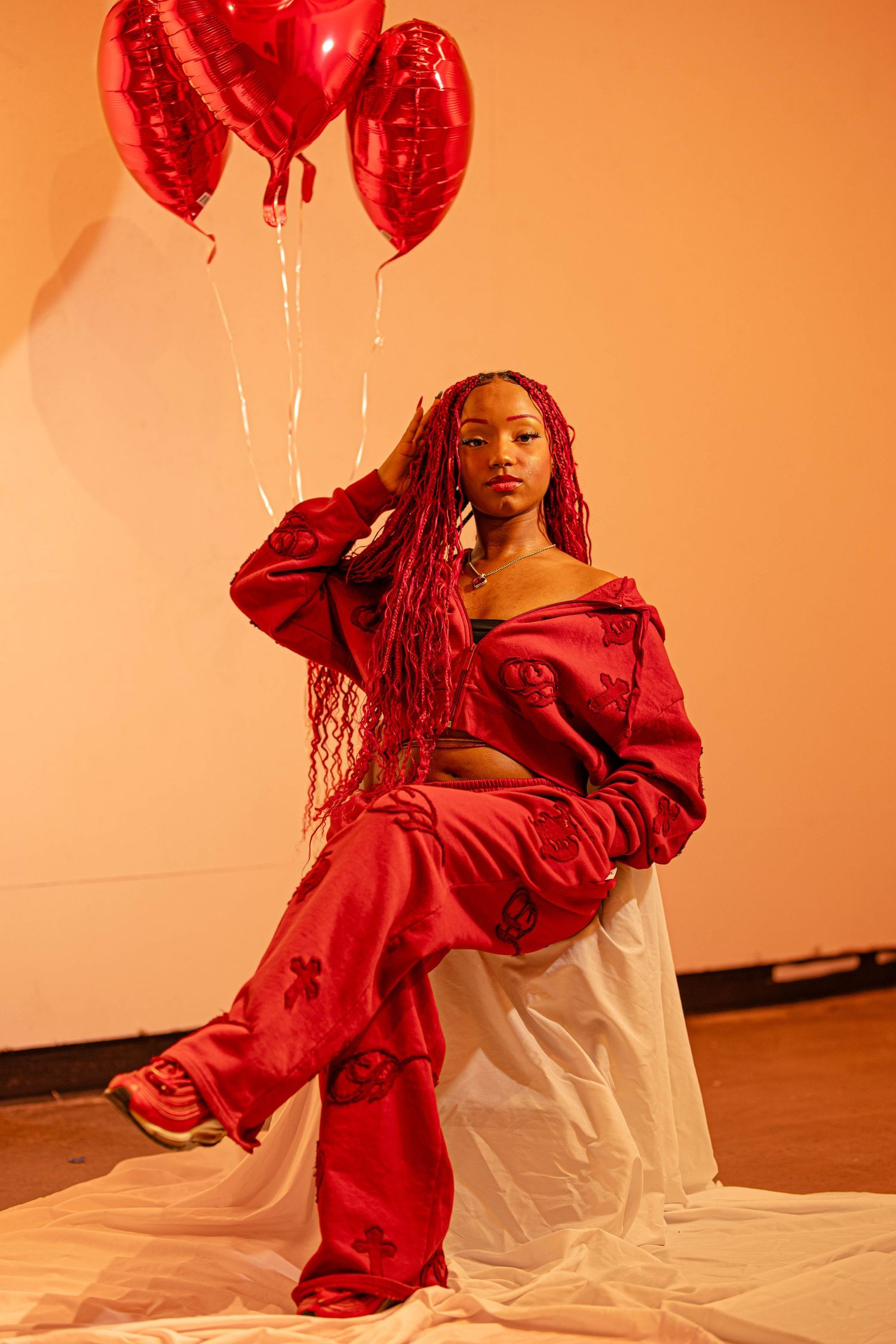 Young woman in red satin outfit holding red heart balloons poses on white fabric in orange-lit studio.