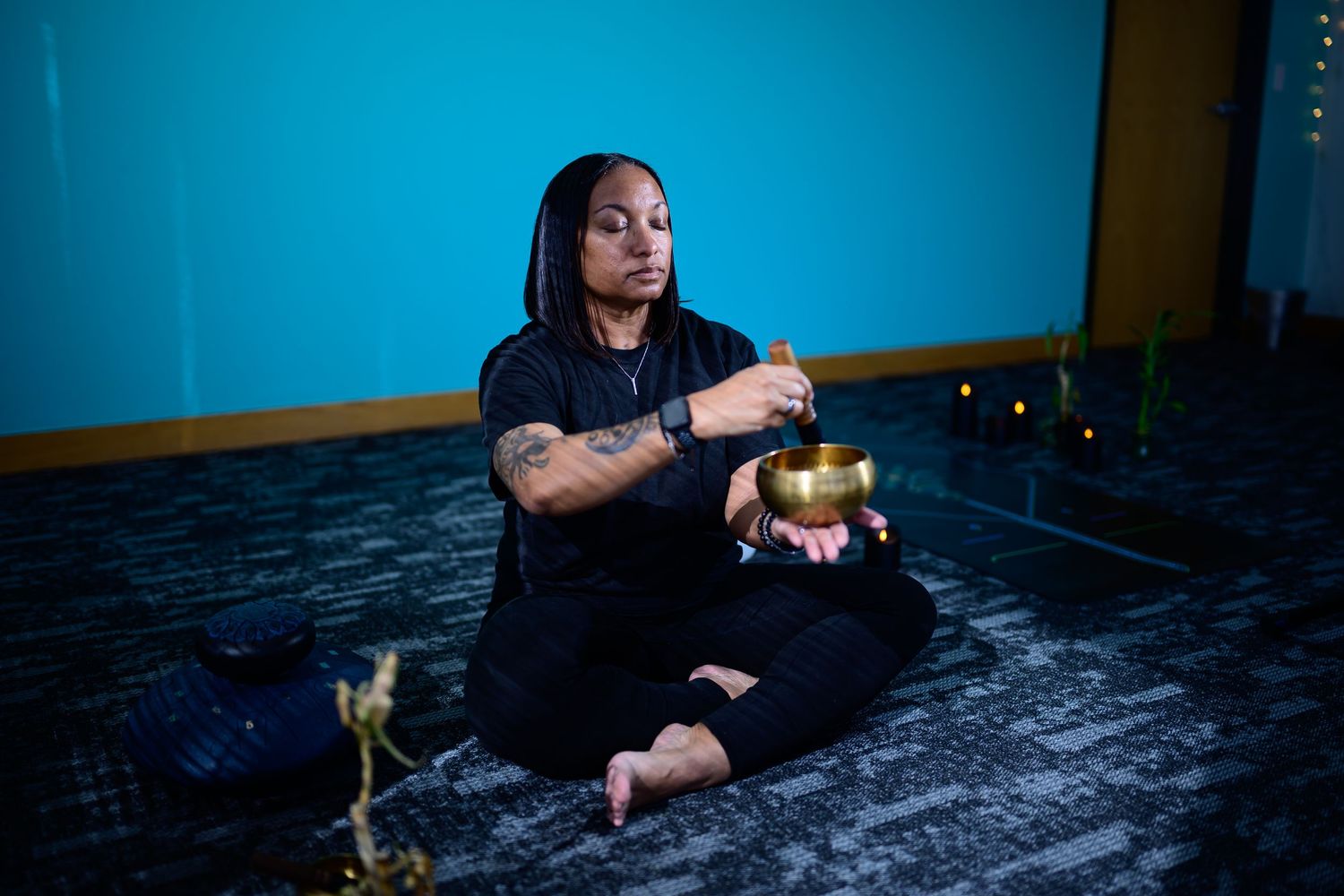 A tattooed woman in all black sits cross-legged on the floor playing a singing bowl in a dimly lit blue room.