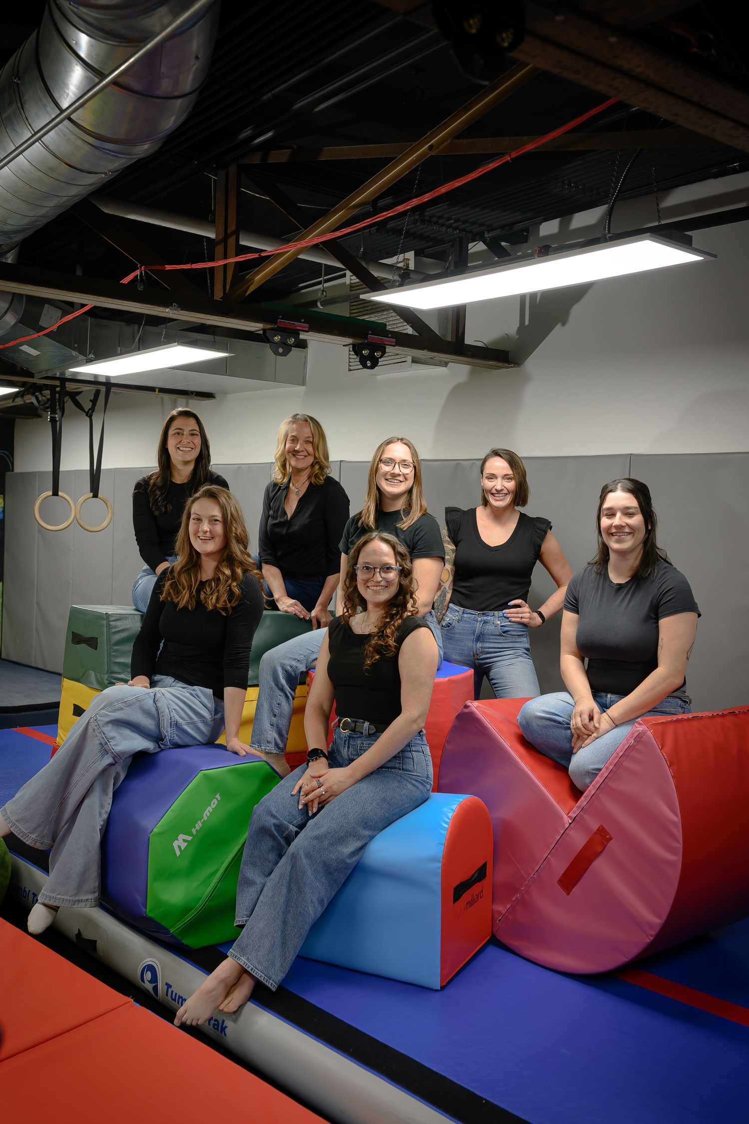 Team of eight women posing on colorful foam equipment in pediatric therapy gym.