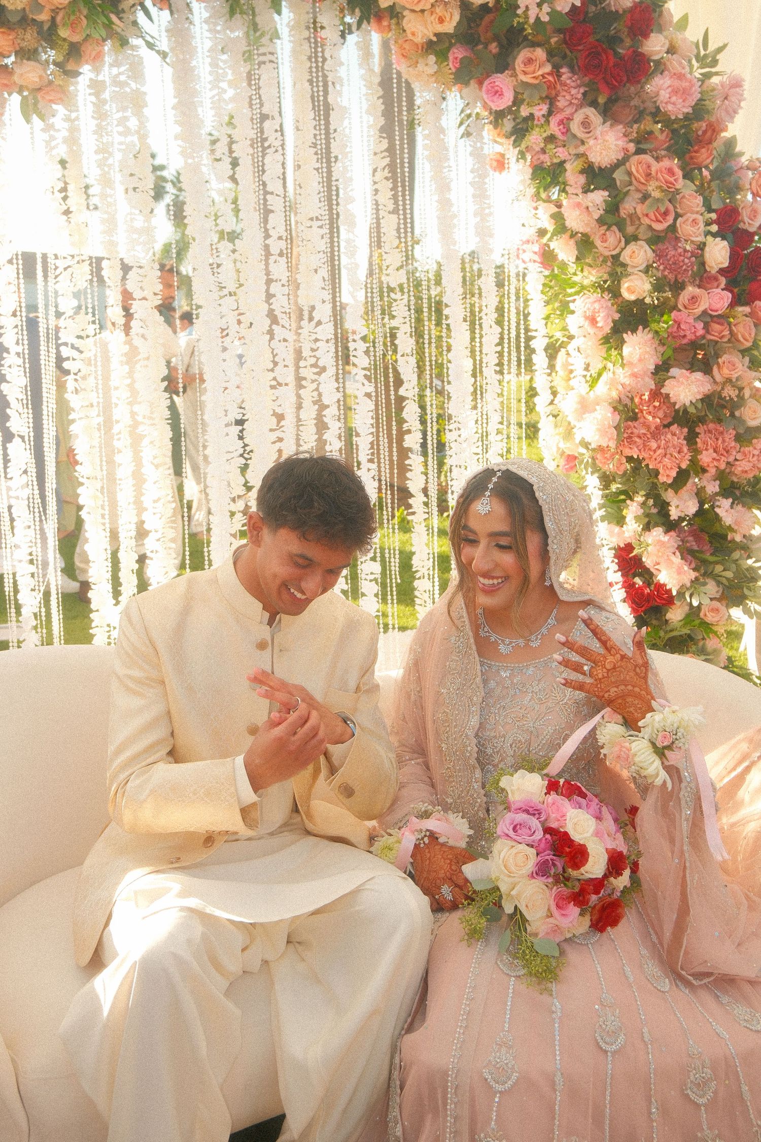 South Asian bride and groom in traditional attire admire wedding rings, seated under floral backdrop.