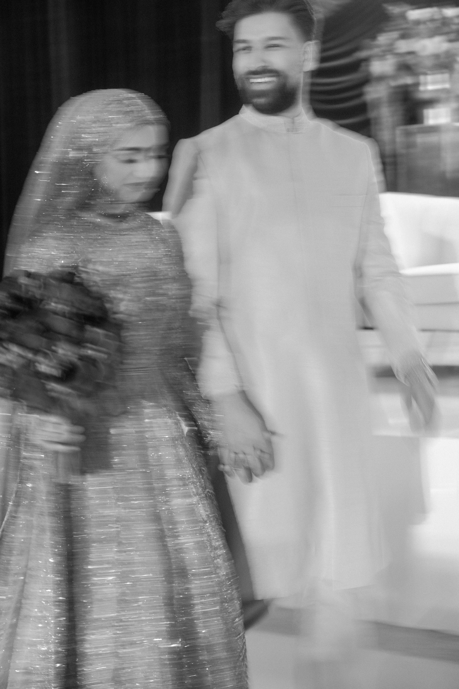 Black and white photo of a smiling groom in kurta holding hands with bride in hijab and gown holding bouquet.