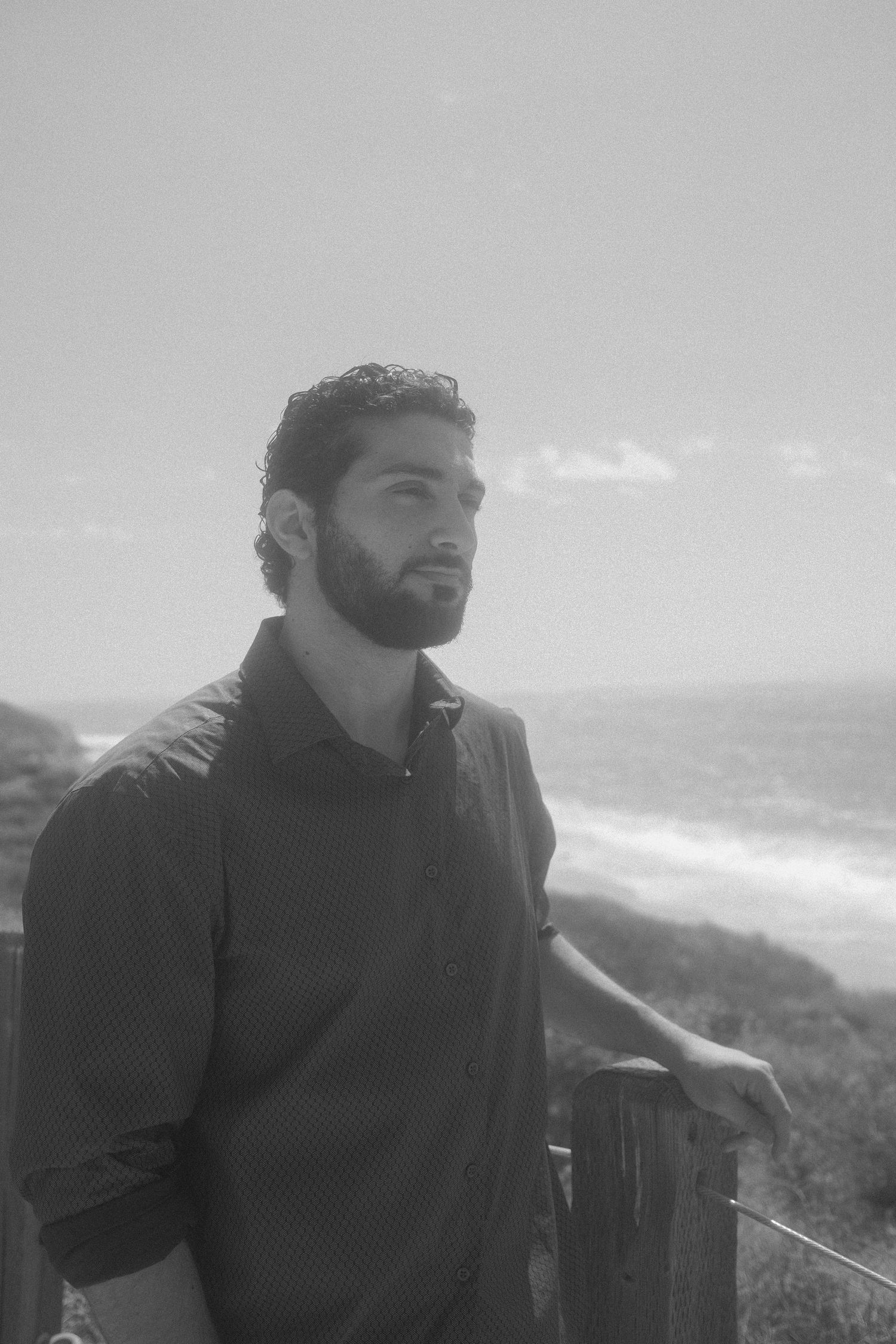 Black and white portrait of a bearded man leaning on a wooden fence overlooking a scenic ocean coastline.