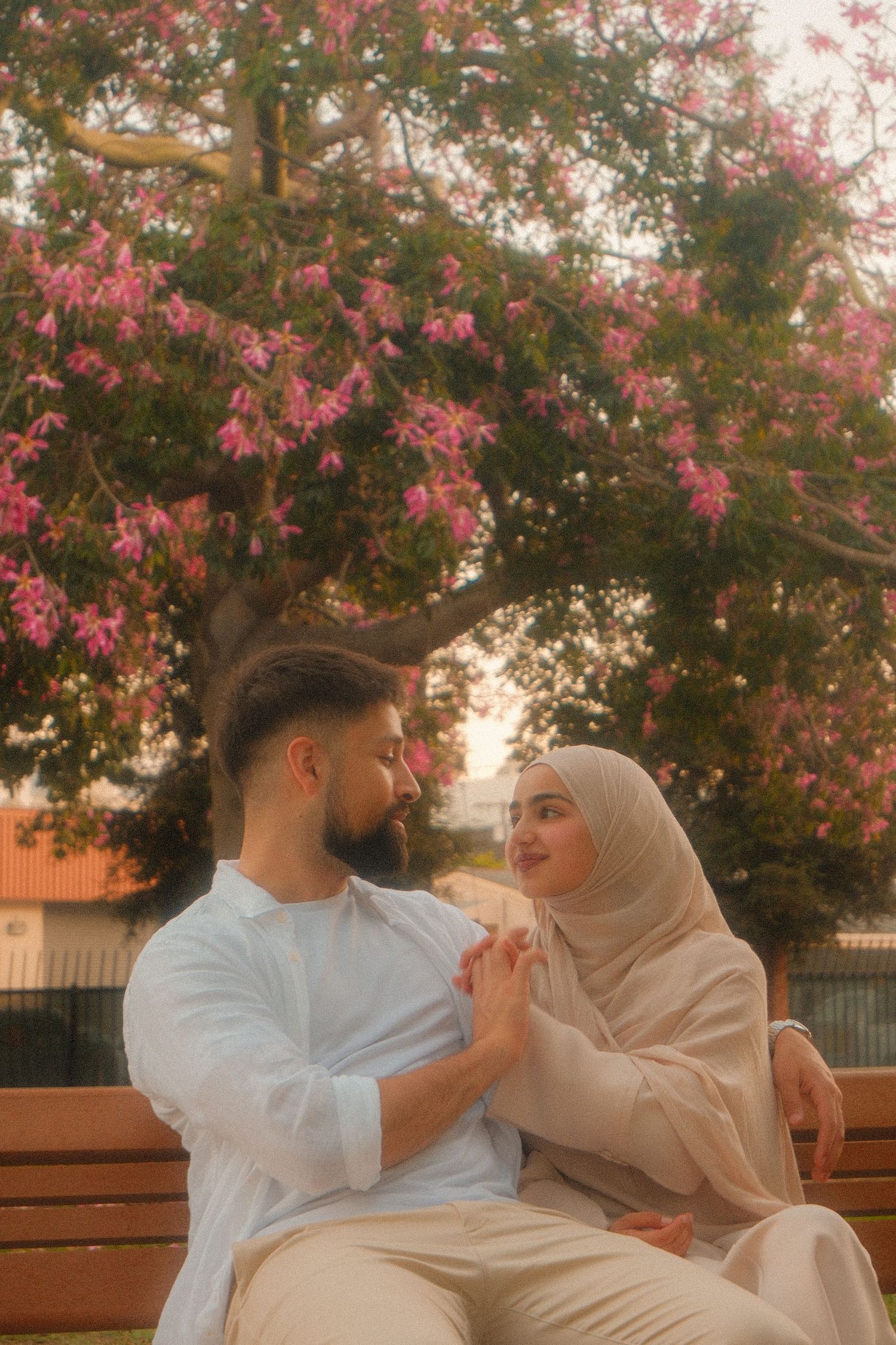 Muslim couple in white and beige attire sit on bench holding hands under pink flowering tree at dusk.