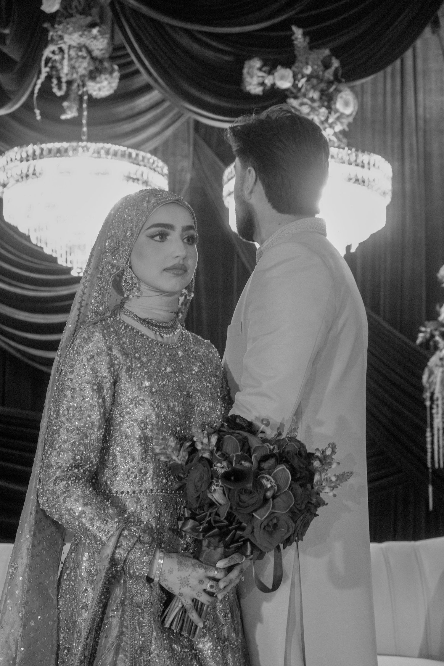 Black and white portrait of Muslim bride in embroidered lehenga holding bouquet with groom at wedding ceremony.