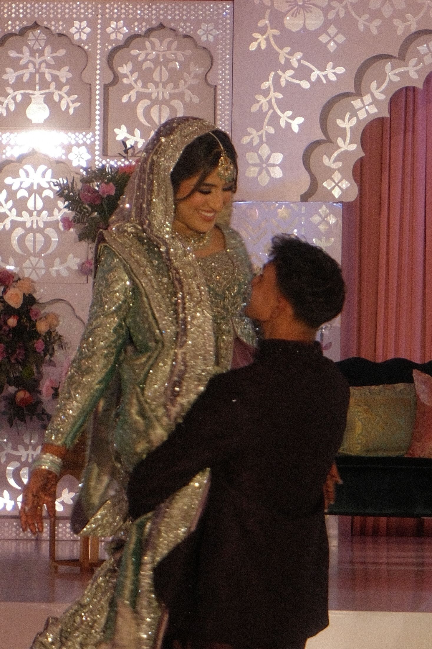 Bride in sparkling green lehenga smiling at groom in black sherwani at decorated wedding stage.