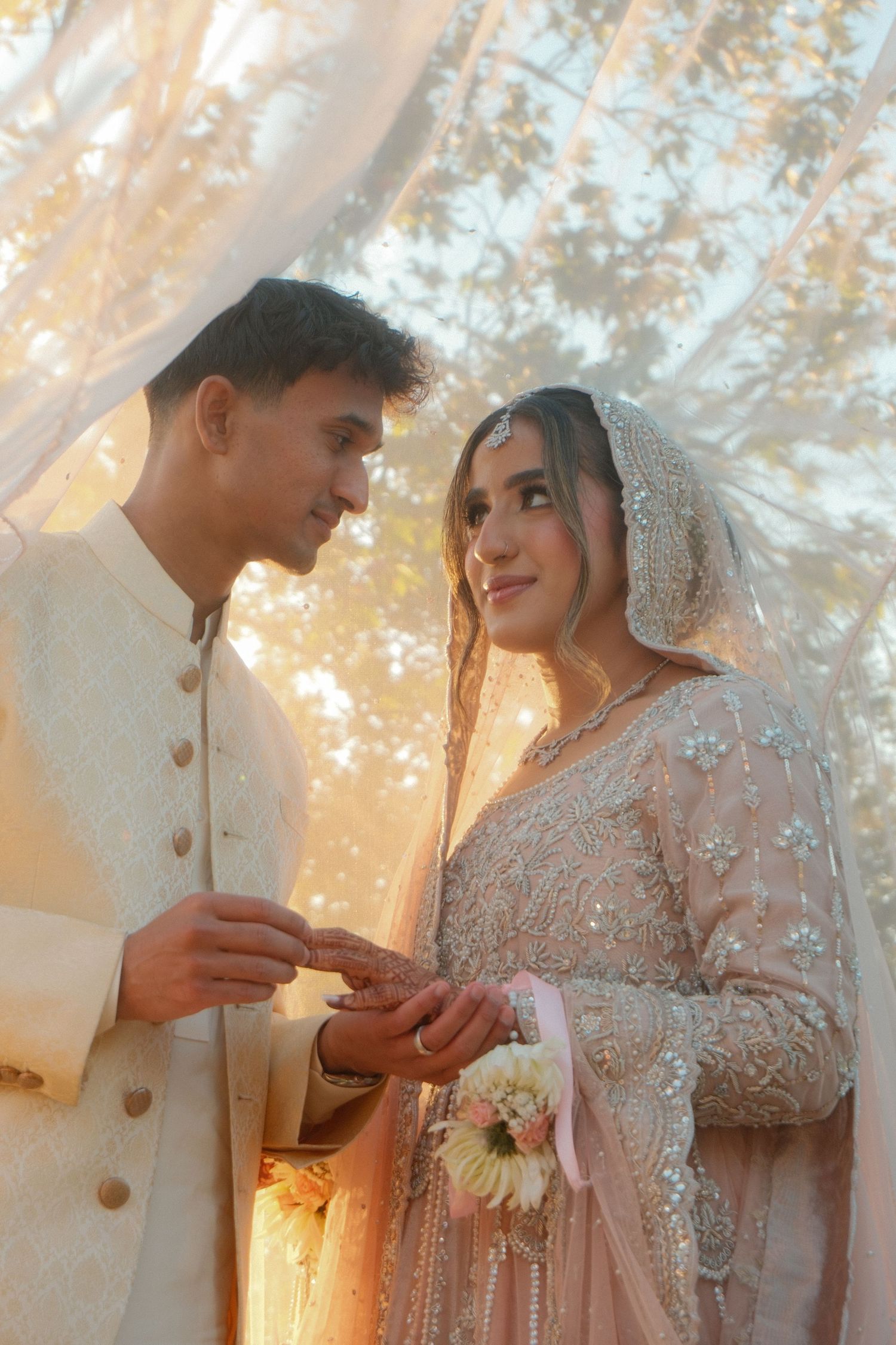 A couple in traditional South Asian wedding attire exchange rings outdoors under soft golden light.