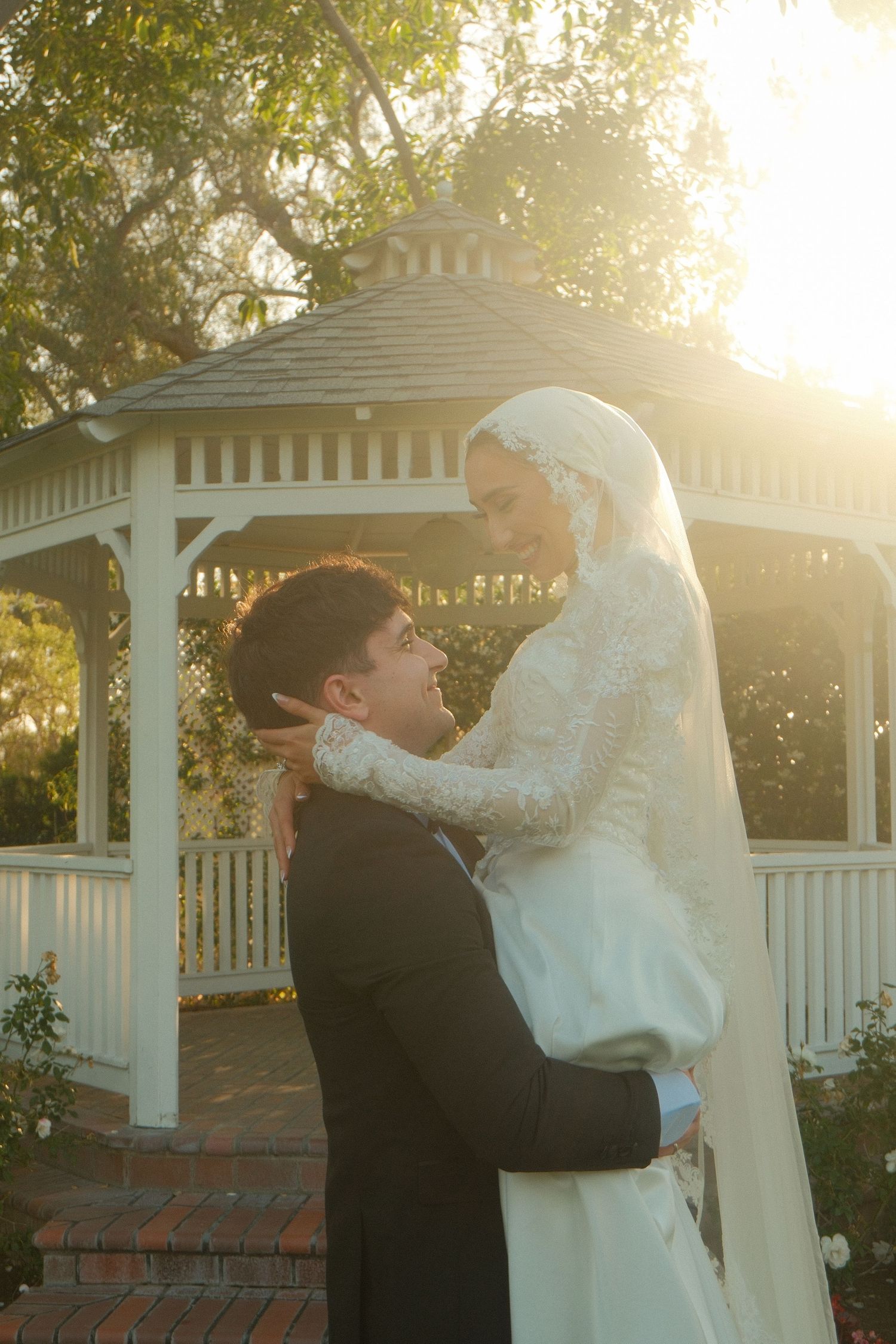 Groom lifts smiling bride in lace wedding dress near white gazebo in golden sunlight.