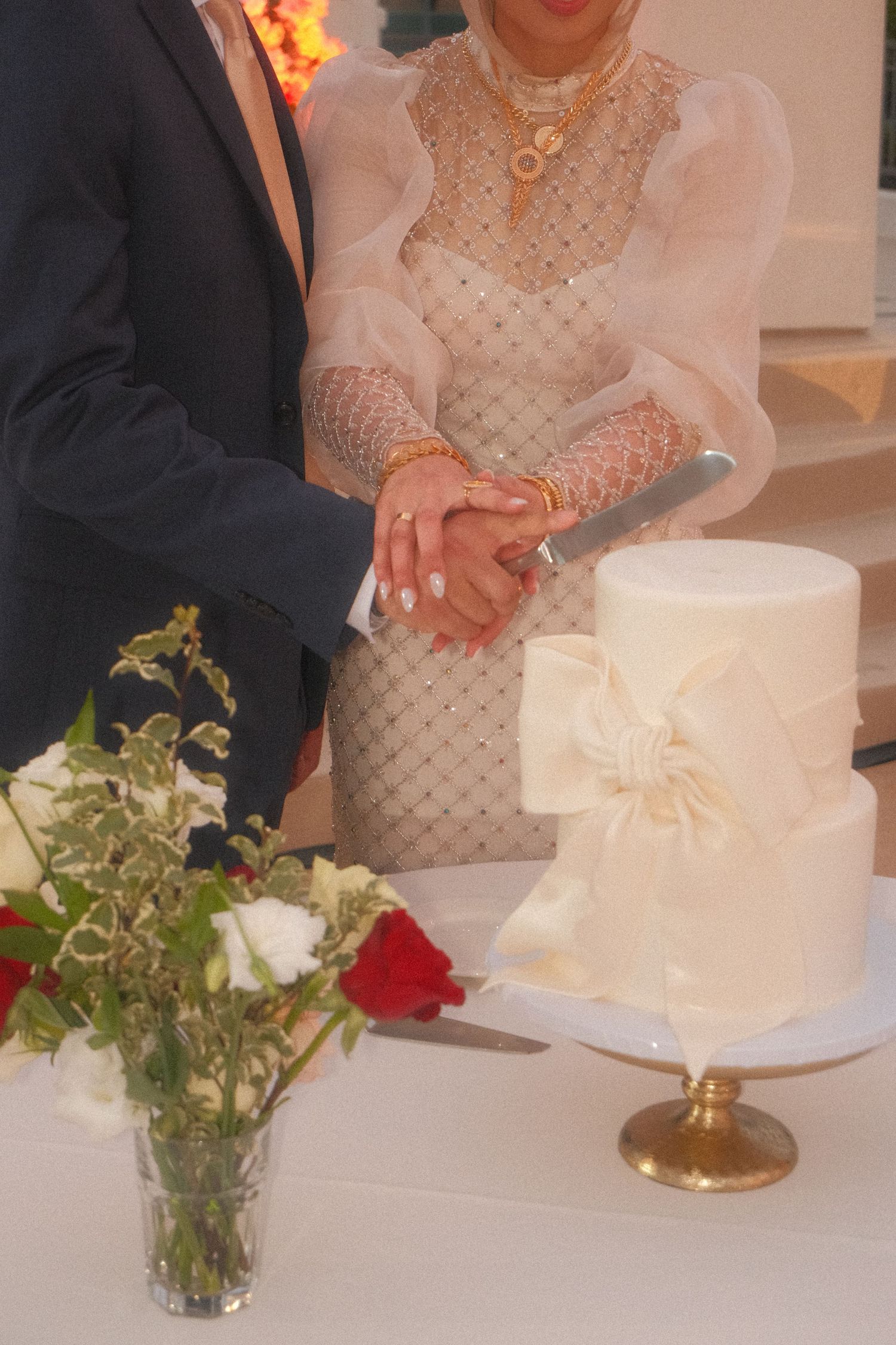 Bride and groom cutting a white two-tier wedding cake decorated with a bow beside red and white flowers.