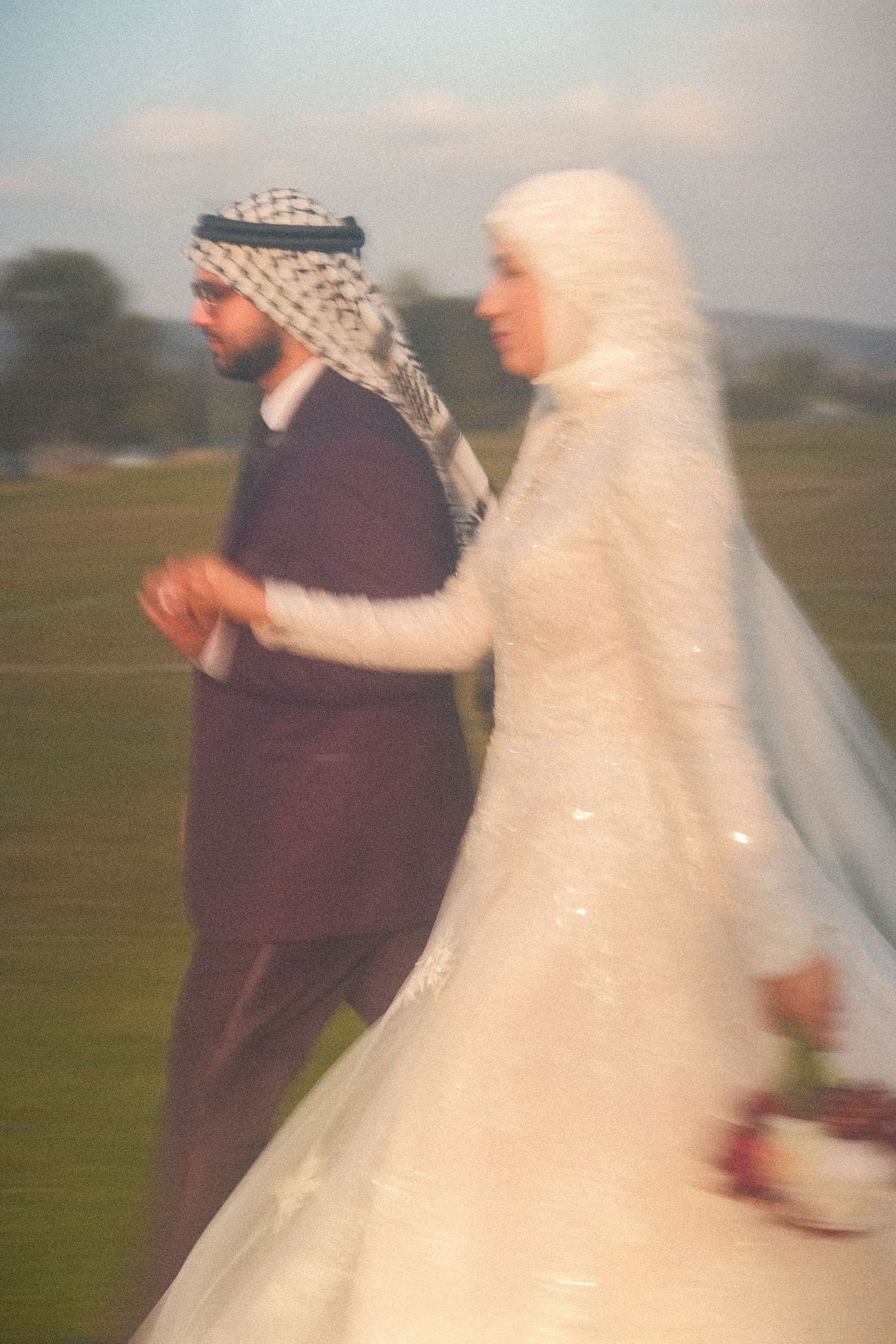 Blurred film photo of Muslim bride in white hijab and groom in maroon walking in a green field.
