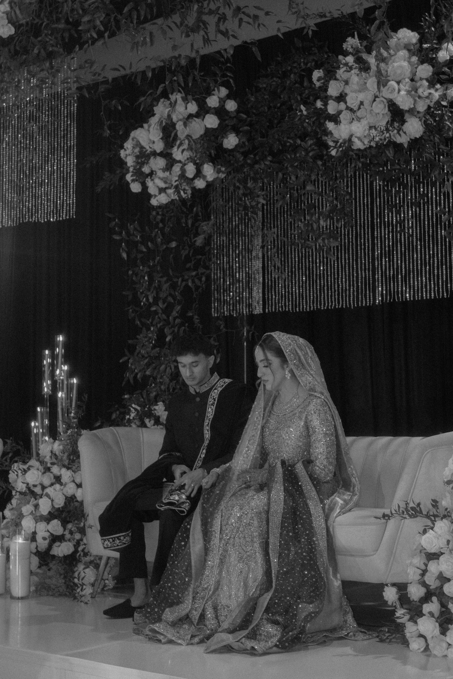 Black and white photo of South Asian wedding couple seated on sofa under floral arch with candles and flower arrangements.