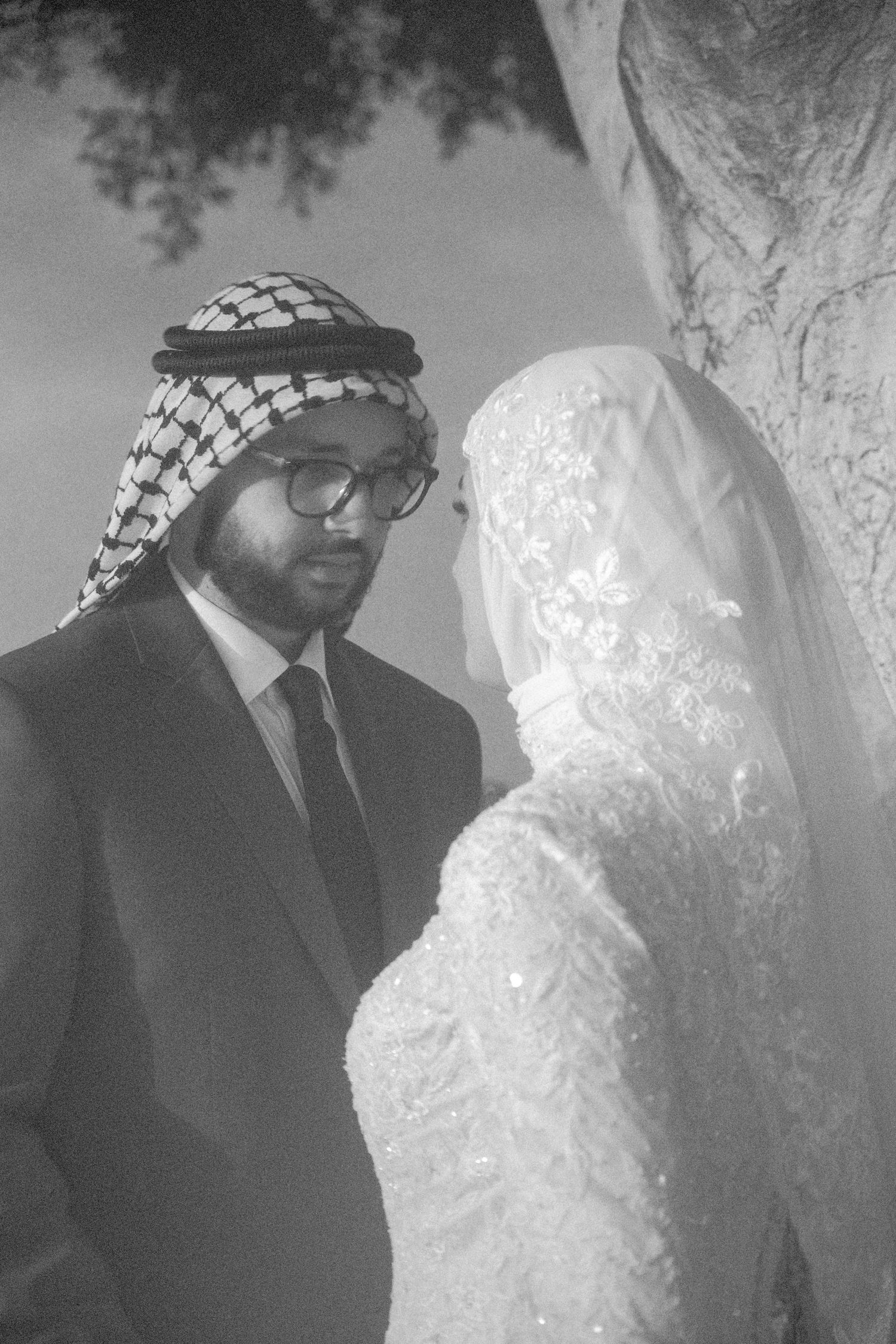 Black and white portrait of a groom in keffiyeh and glasses facing his veiled bride in a lace gown.