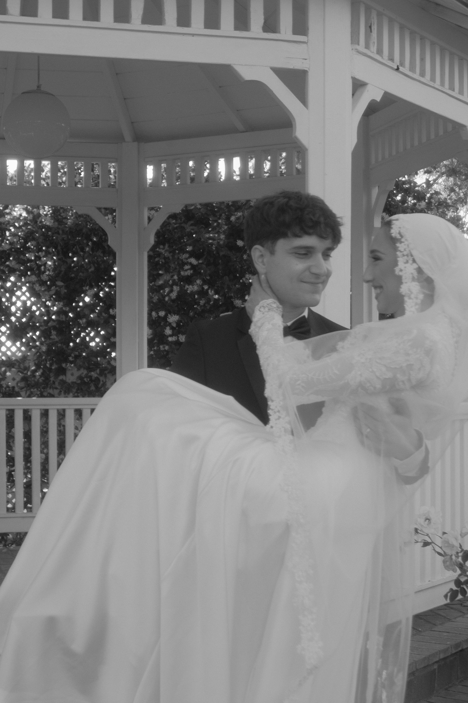 Black and white photo of groom carrying bride in white gown near wedding gazebo.