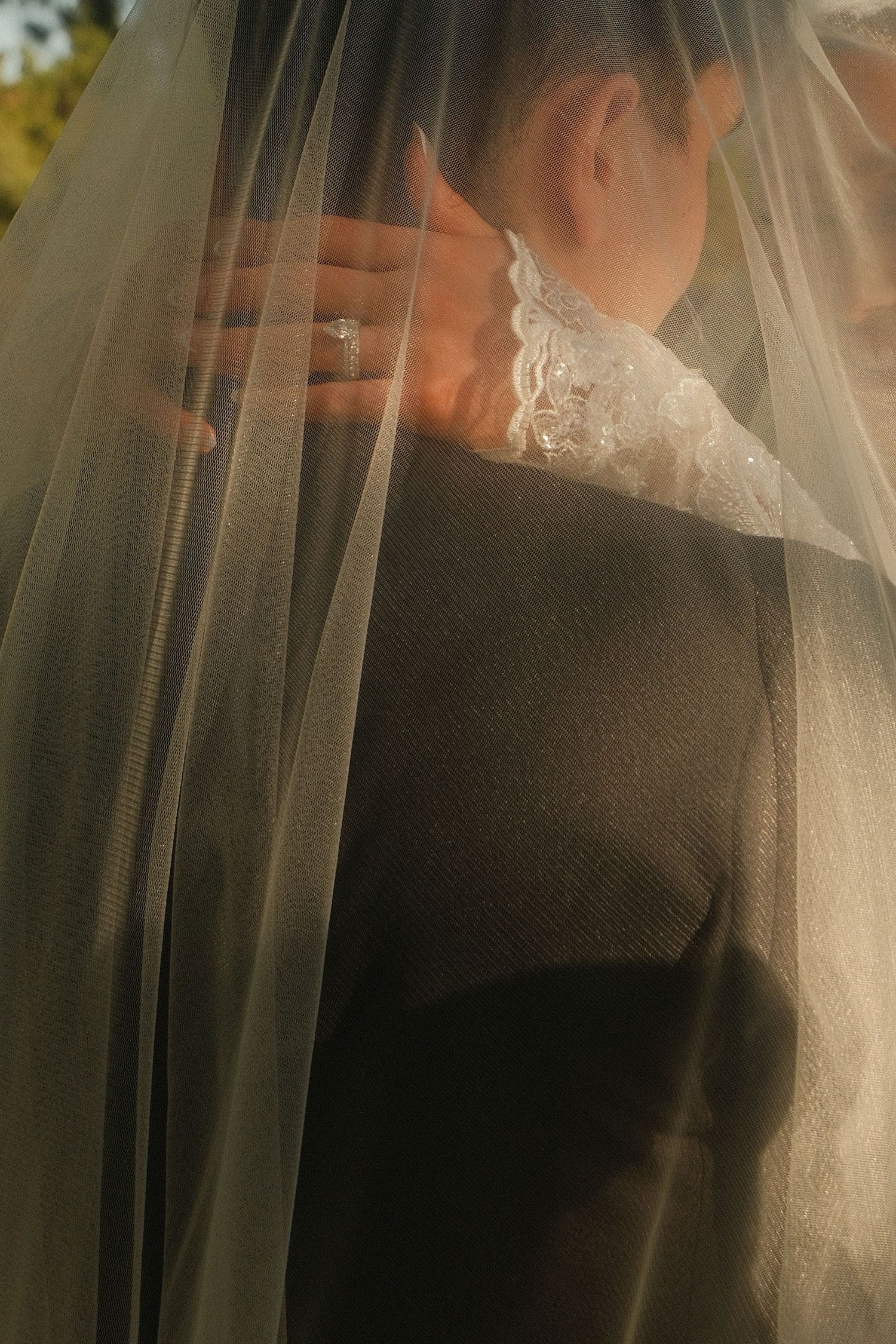 Close-up of bride's hand with diamond ring resting on groom's neck beneath sheer lace veil.