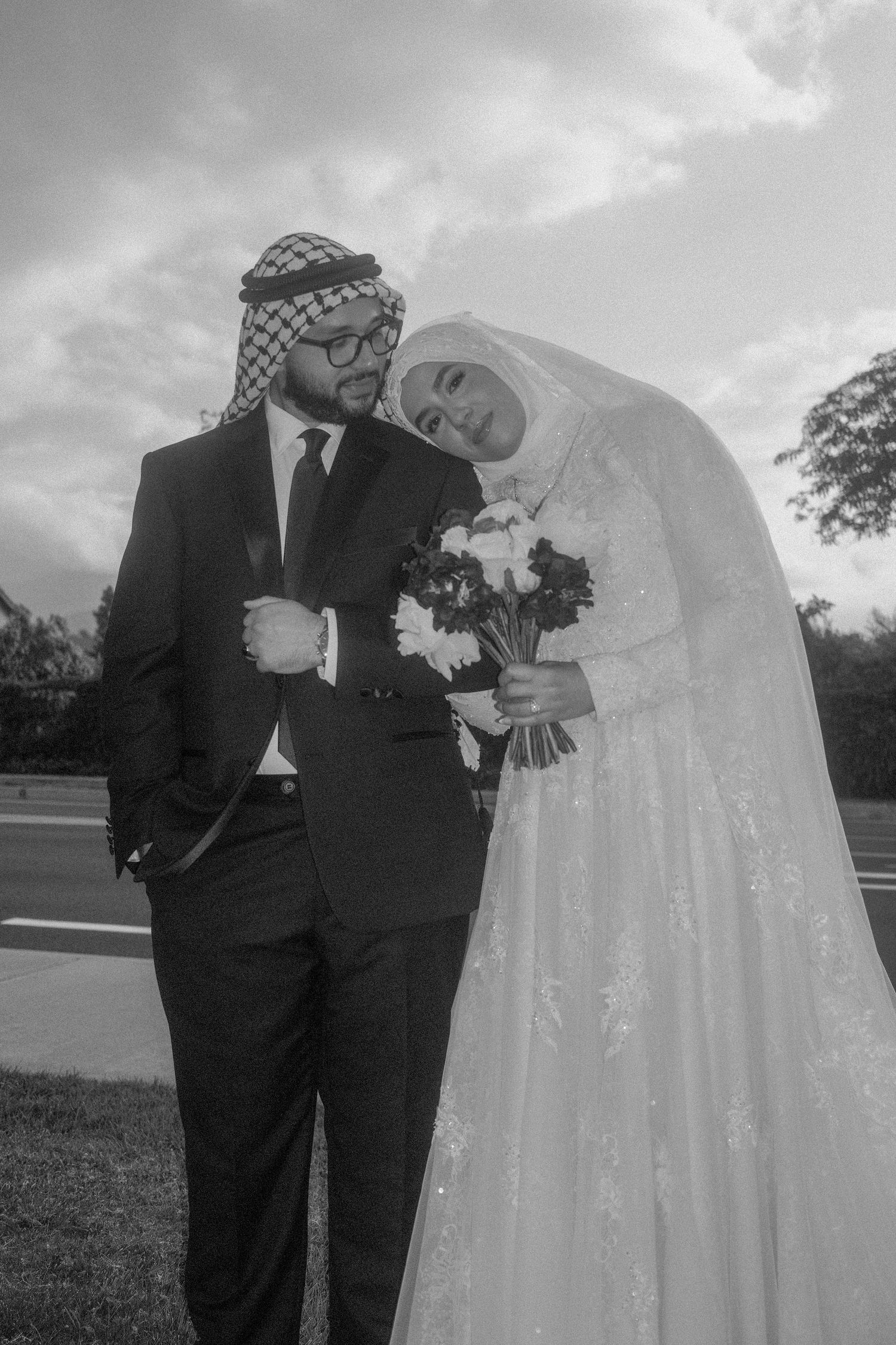 Black and white portrait of Muslim bride leaning on groom in suit and keffiyeh, bride holding bouquet.