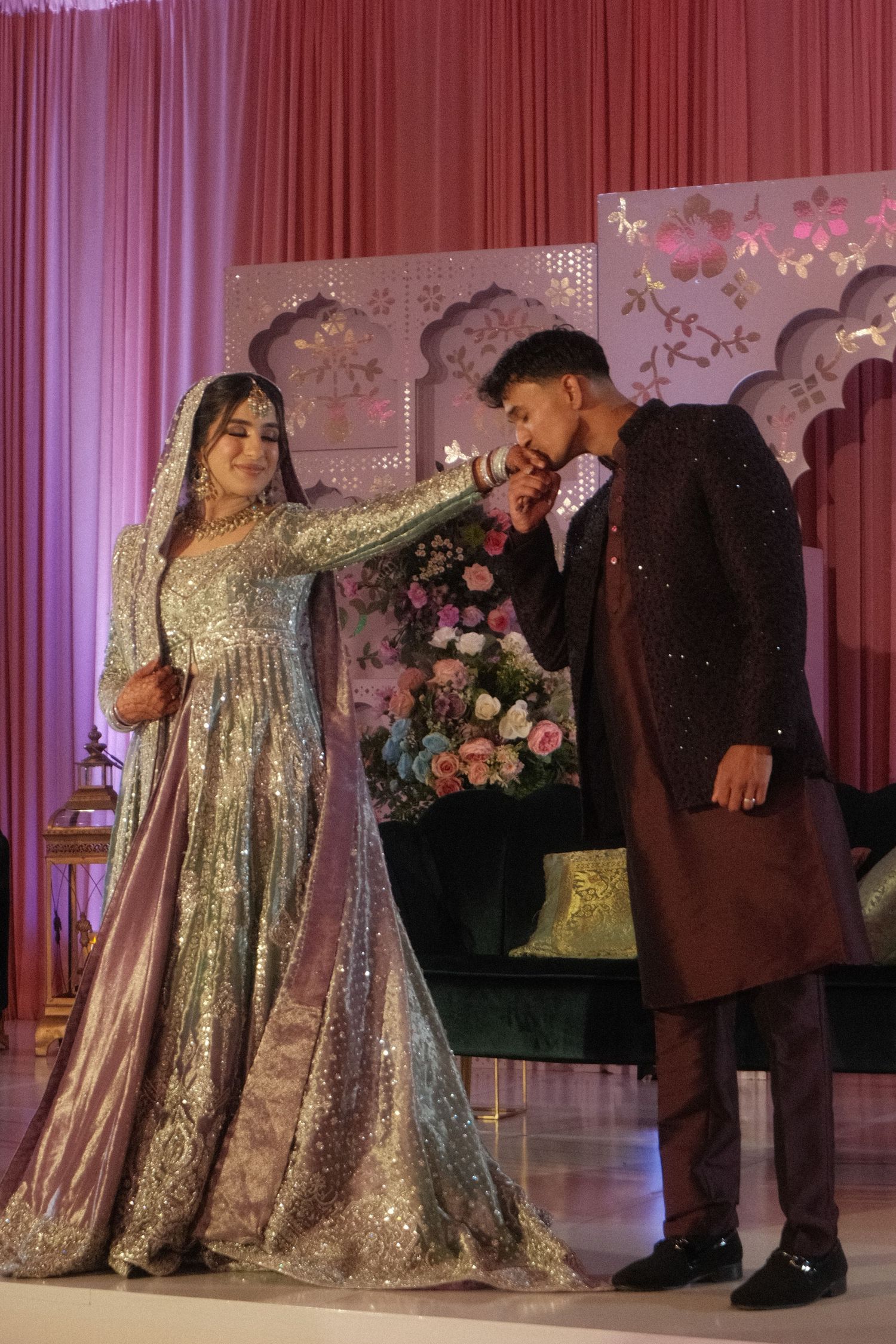 Groom in dark sherwani kissing bride's hand at elegant South Asian wedding ceremony on stage.