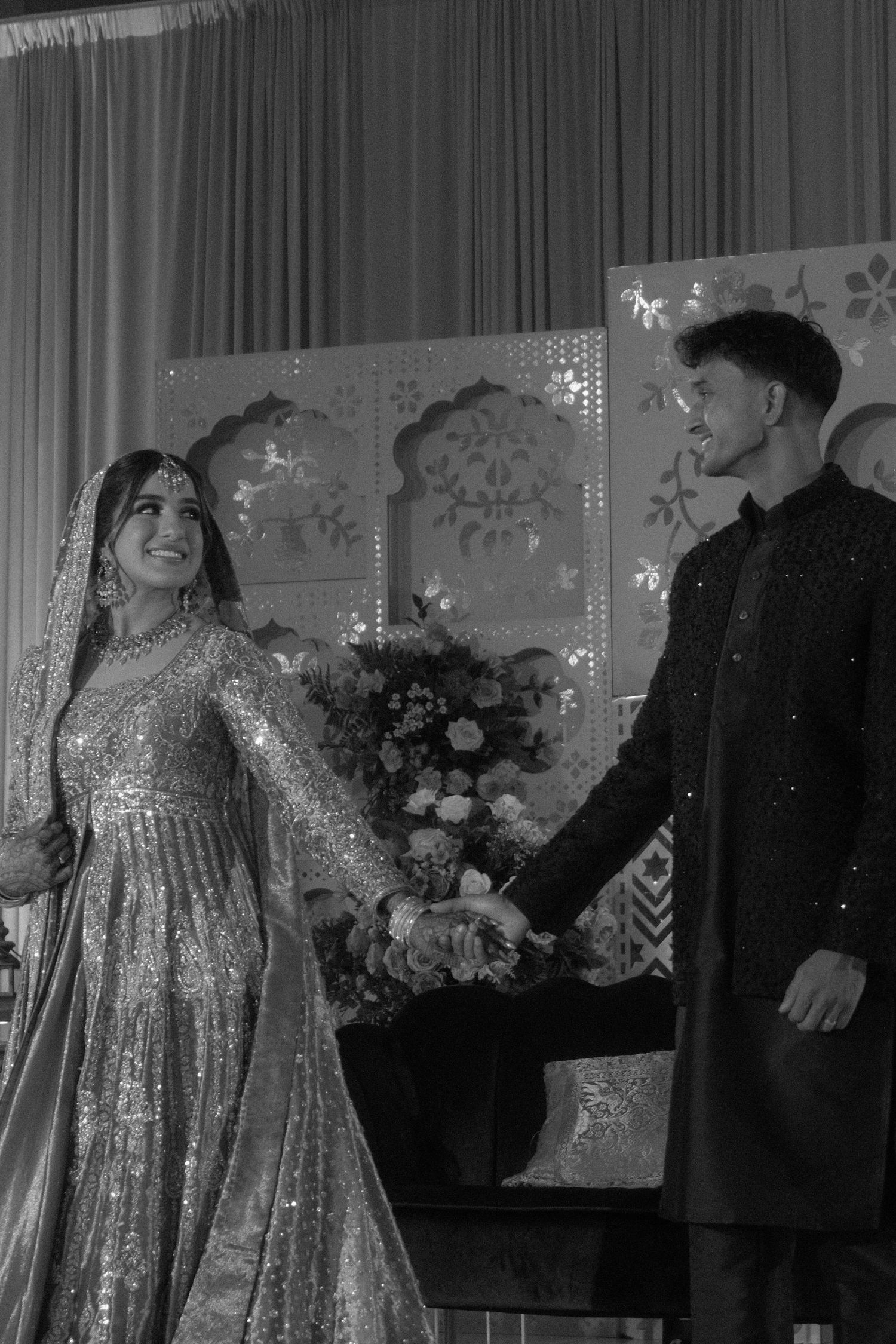Black and white photo of smiling South Asian bride and groom holding hands at wedding reception.