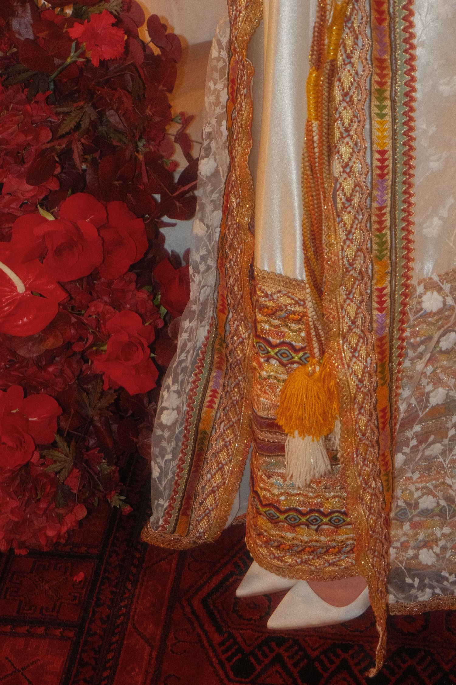 Detail of a traditional embroidered belt with orange tassels beside red floral decorations.