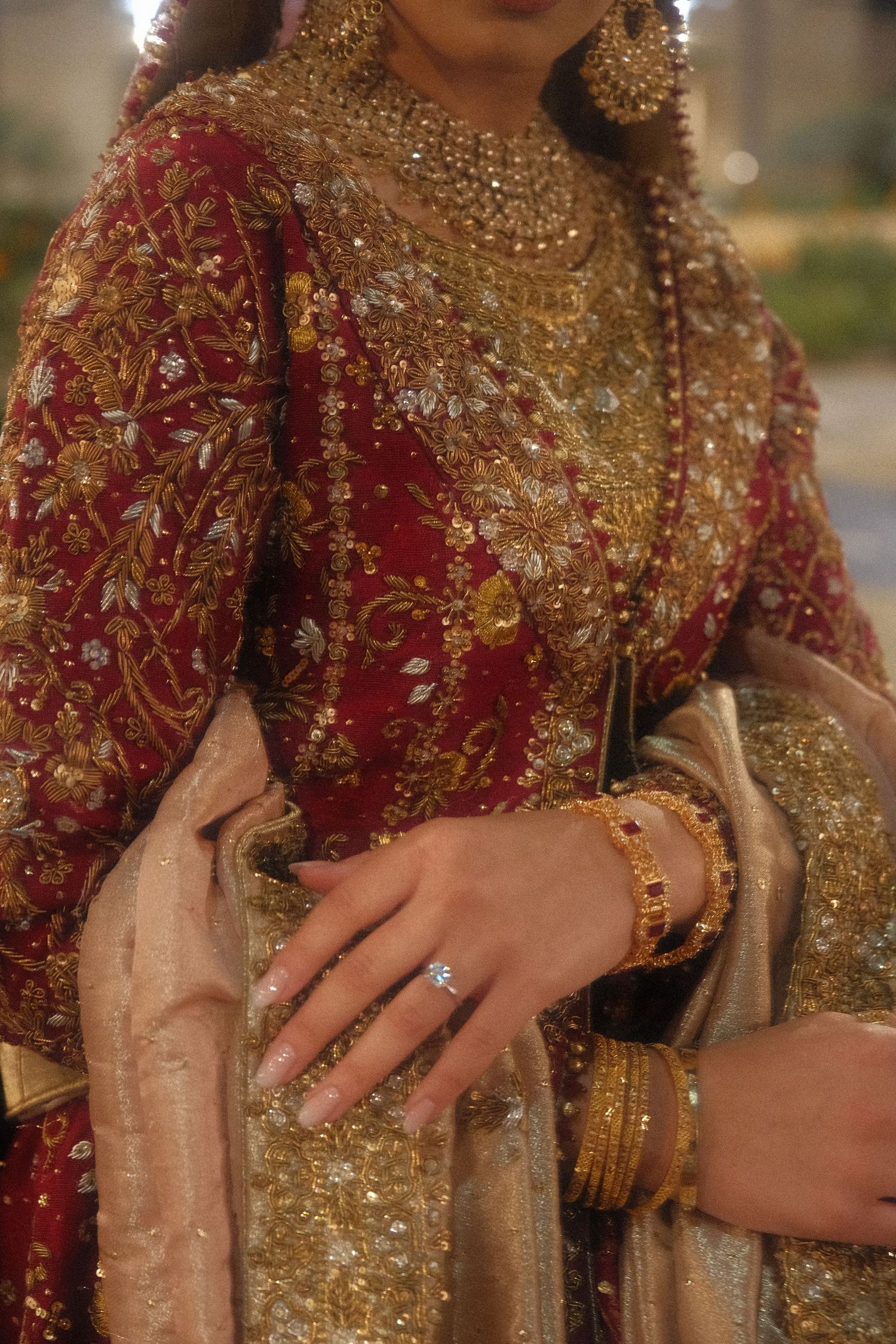 Pakistani bride in red and gold embroidered bridal dress displaying diamond ring and gold bangles.
