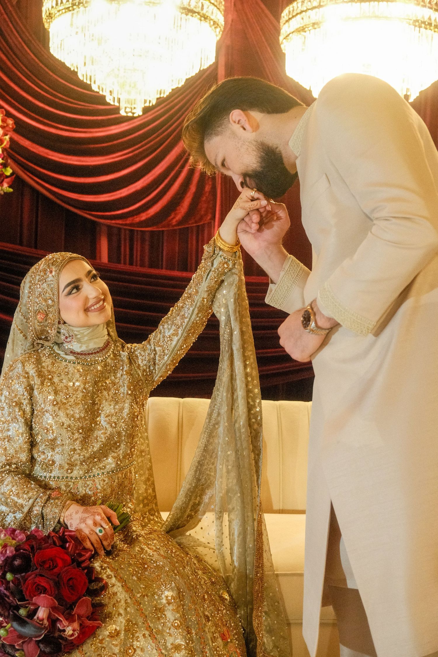Muslim groom in white sherwani kissing bride's hand as she smiles in gold embroidered lehenga with hijab.