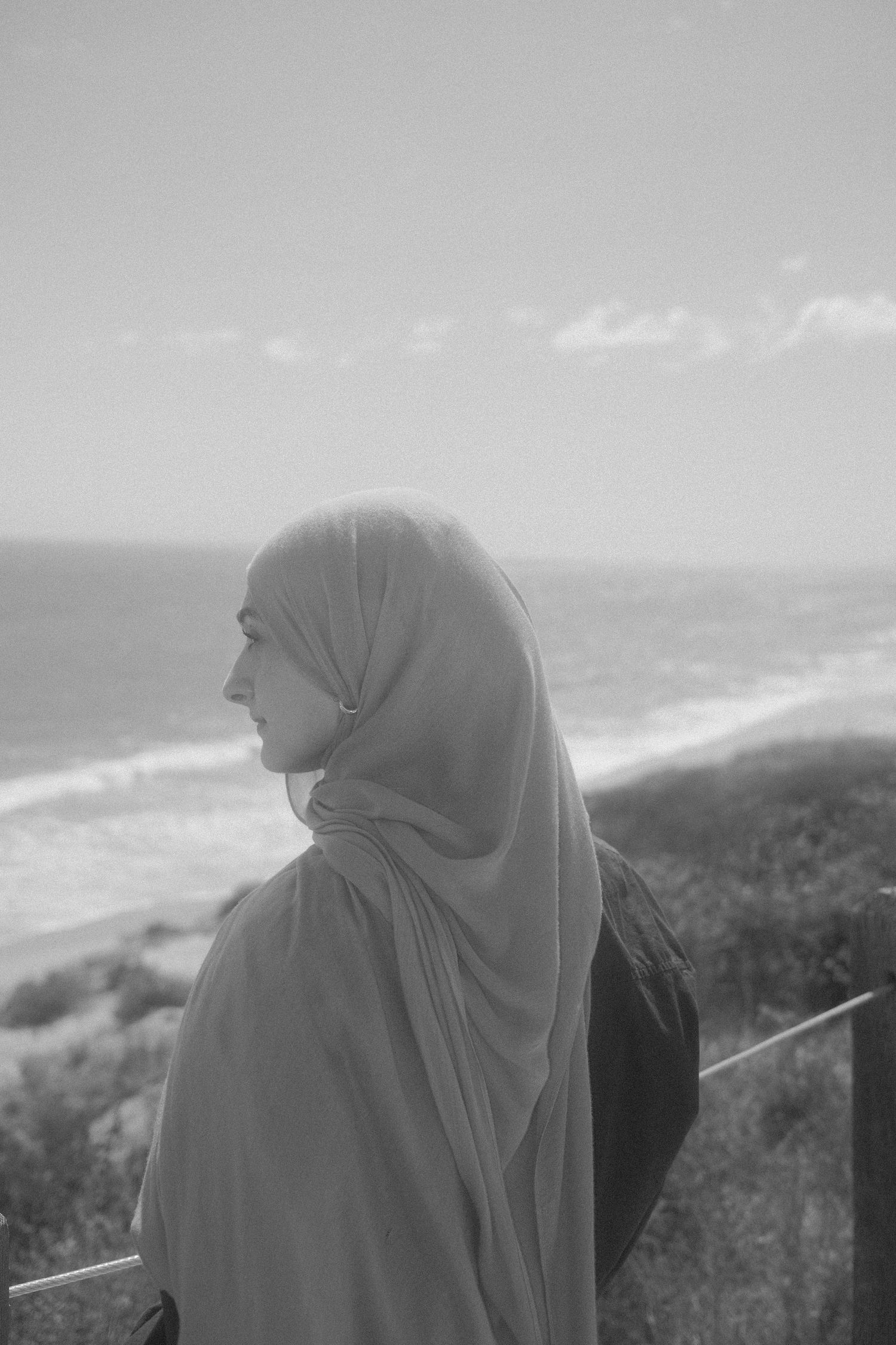 Black and white photo of a woman wearing a hijab overlooking a scenic coastal beach landscape.