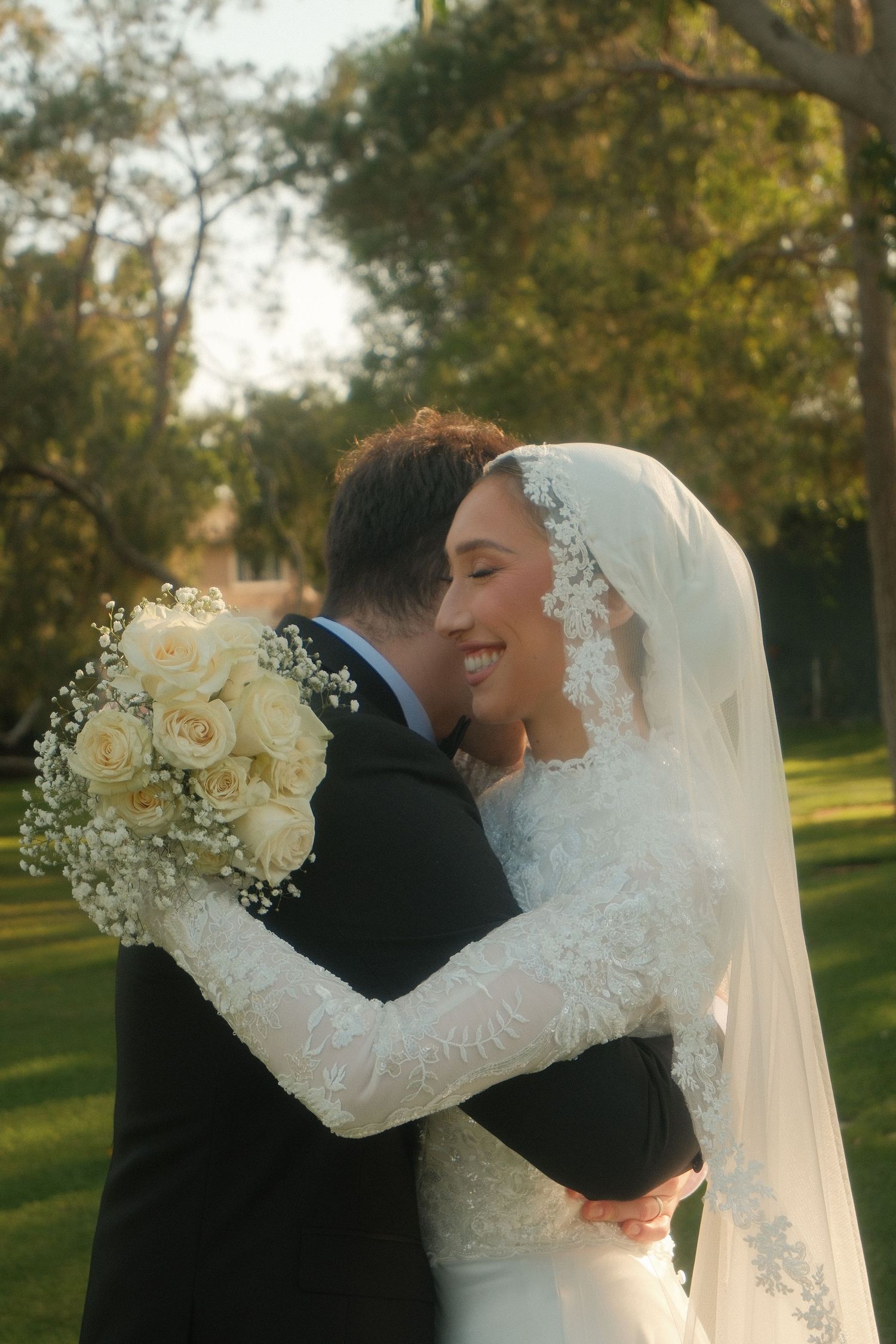 Smiling bride in white lace veil embracing groom holding white rose bouquet in golden sunlit garden.