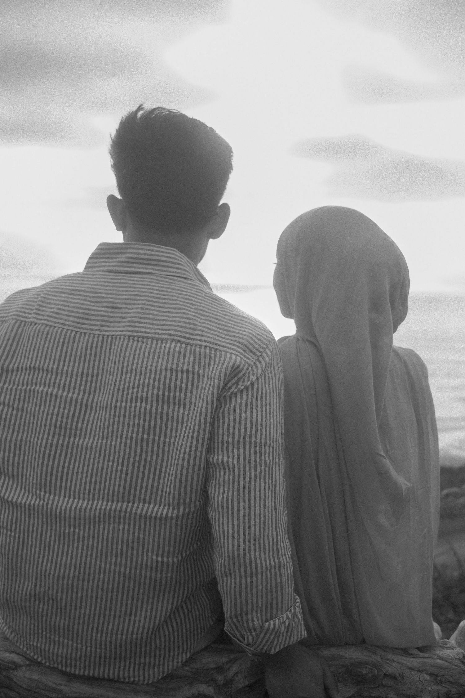 Black and white rear view of a couple sitting on rocks by the ocean, woman wearing a hijab.