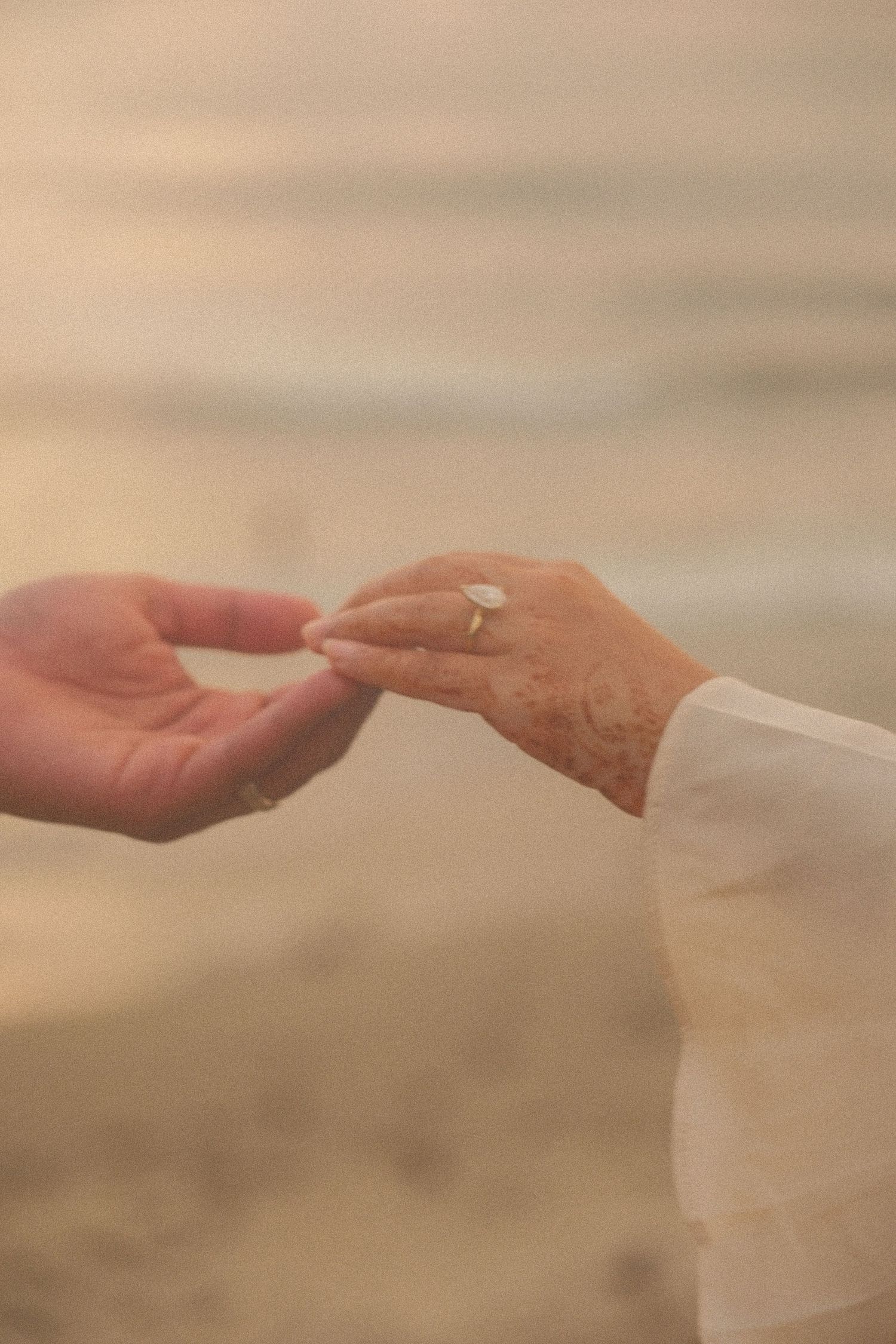 Close-up of an elderly woman's hand with a diamond engagement ring touching a man's hand on a beach.