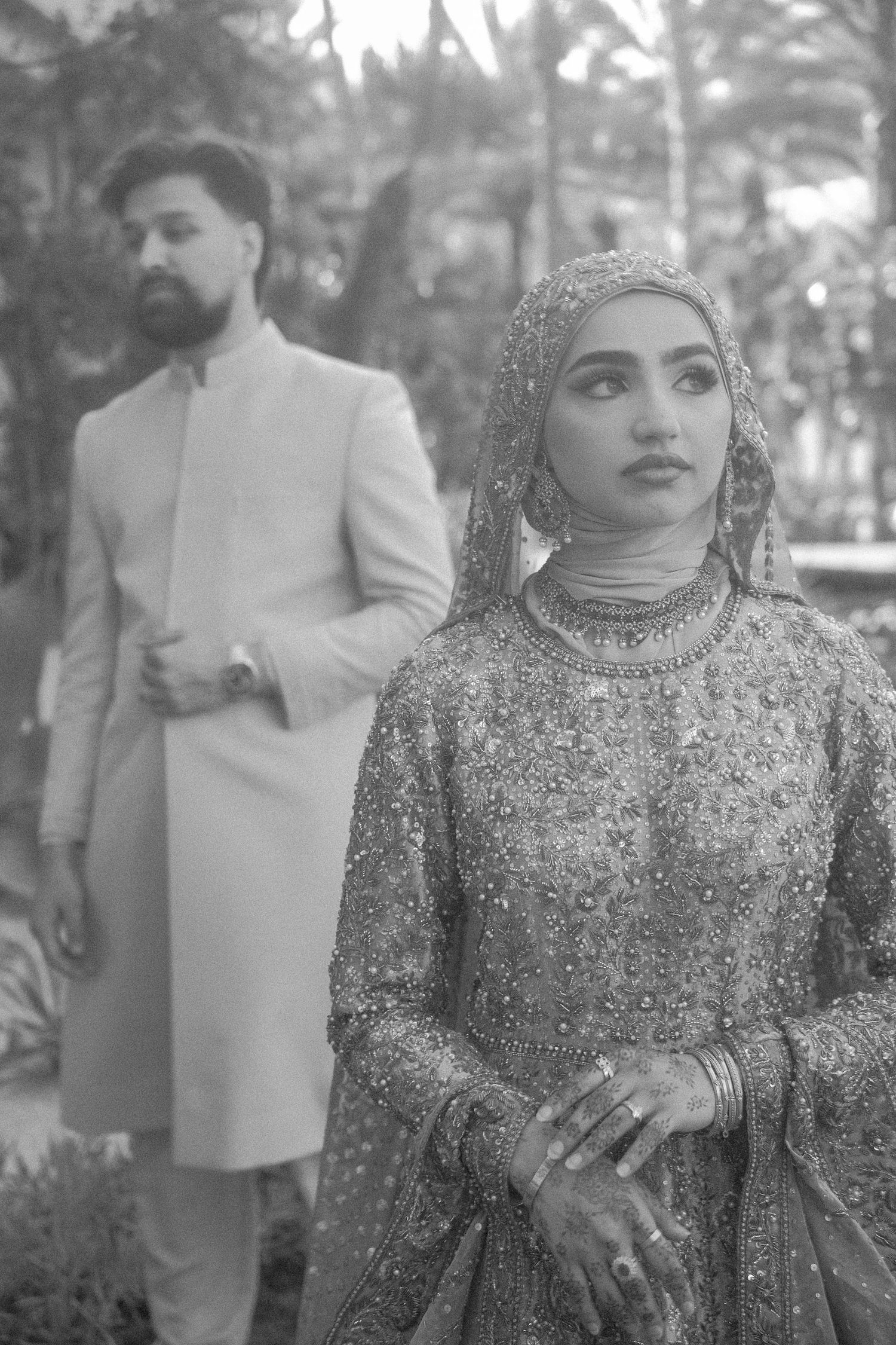 Black and white portrait of Pakistani bride in embellished lehenga with groom in sherwani outdoors.