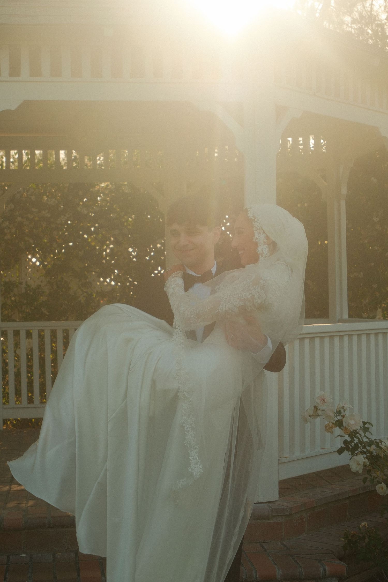 Groom carrying bride in white wedding dress outside sunlit gazebo.