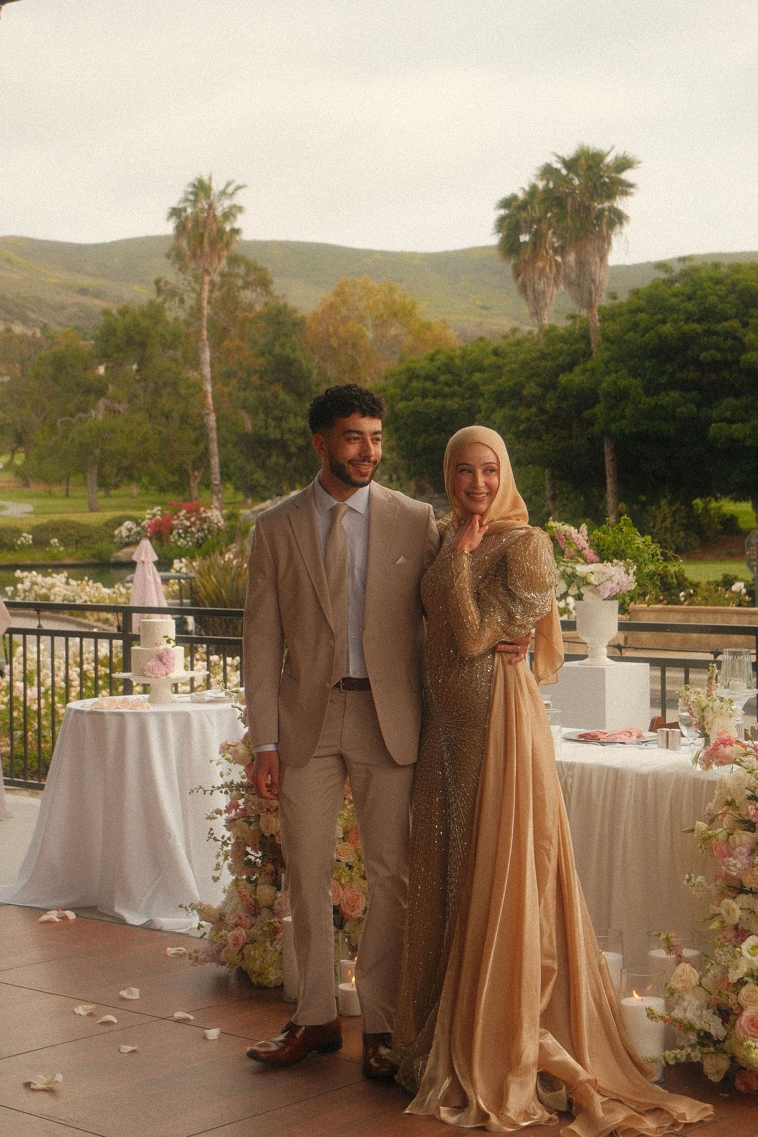 A couple in elegant wedding attire poses outdoors, with palm trees and green hills in the background.