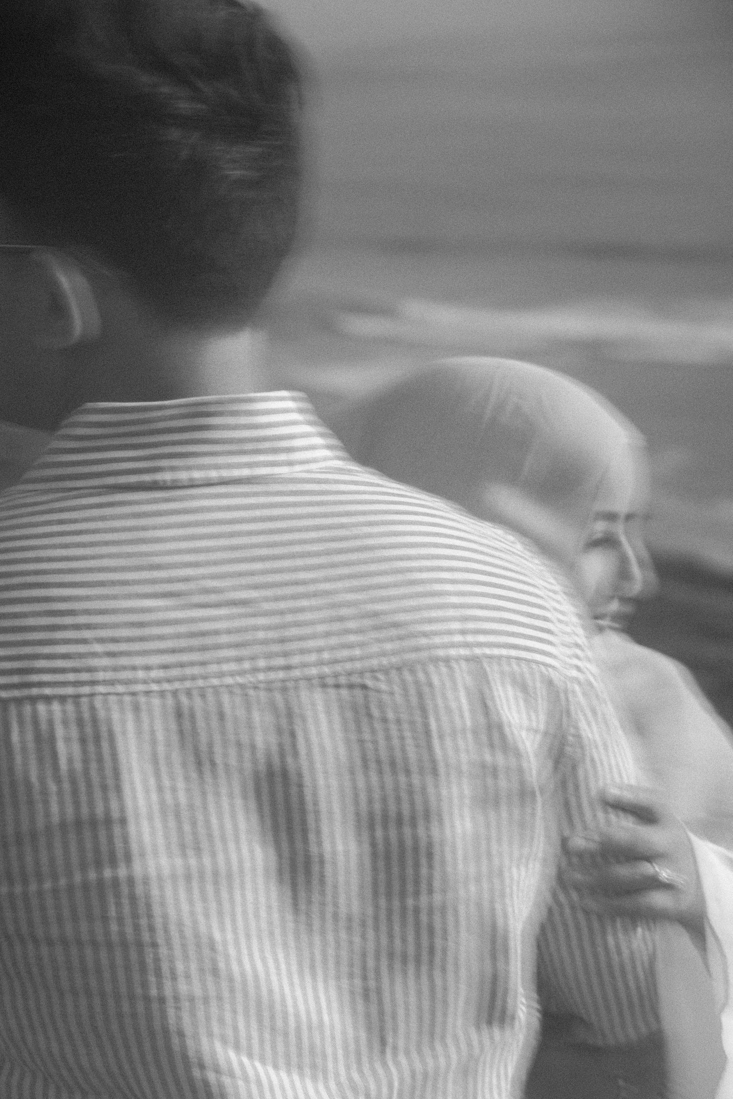 Black and white photo of couple embracing near ocean, man in striped linen shirt with woman beside him.