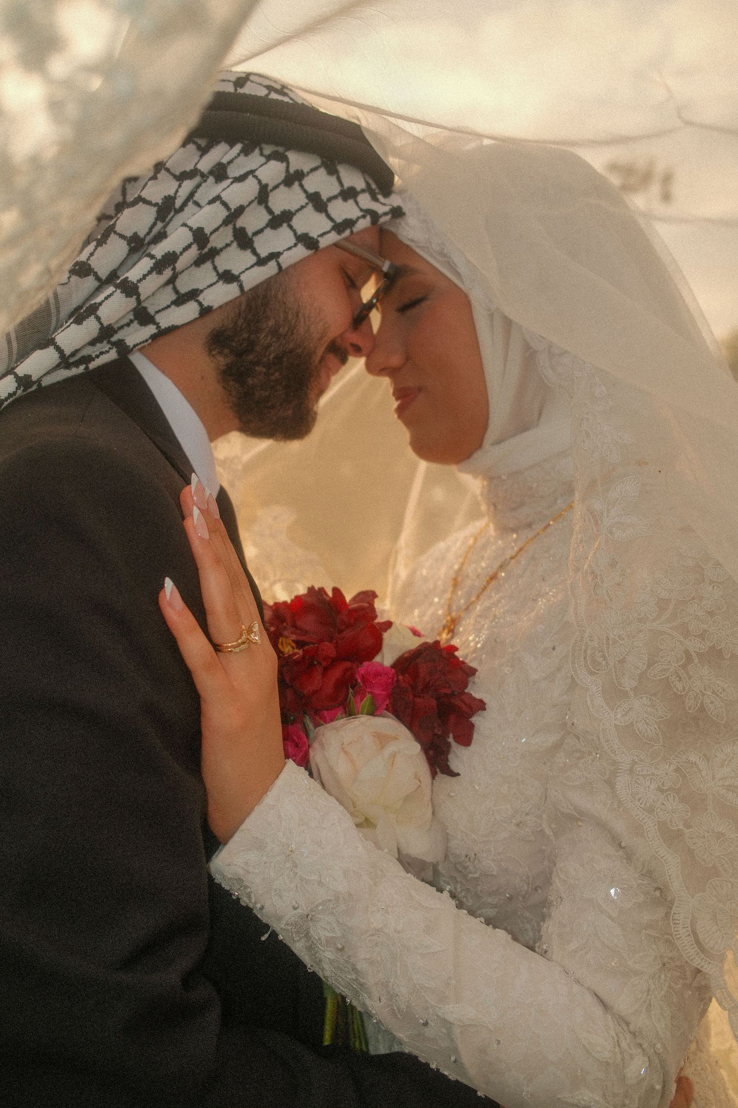 Muslim bride and groom in traditional attire sharing an intimate moment under a veil with red bouquet.