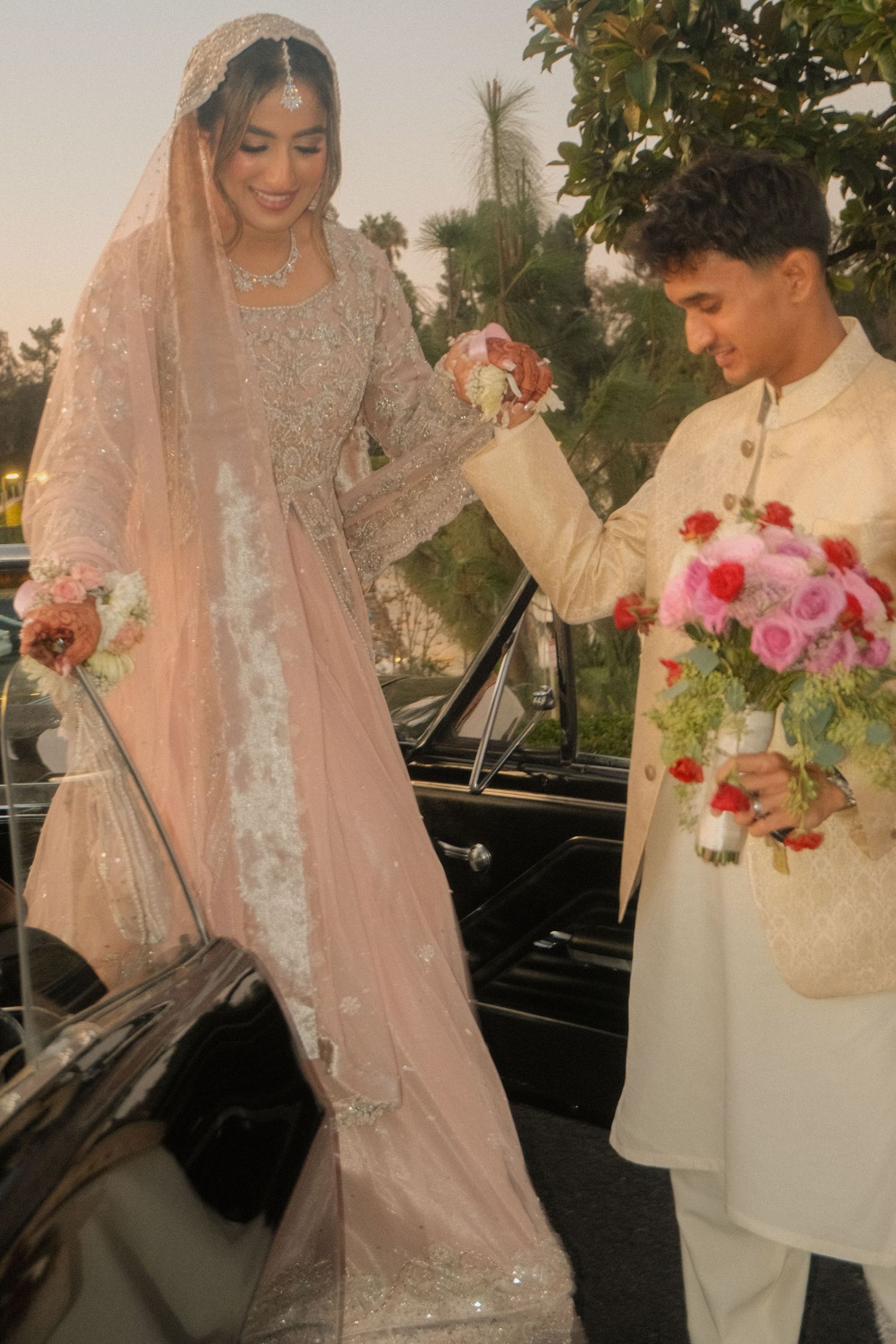 South Asian bride in embroidered pink lehenga with veil helped by groom in cream sherwani near vintage car.