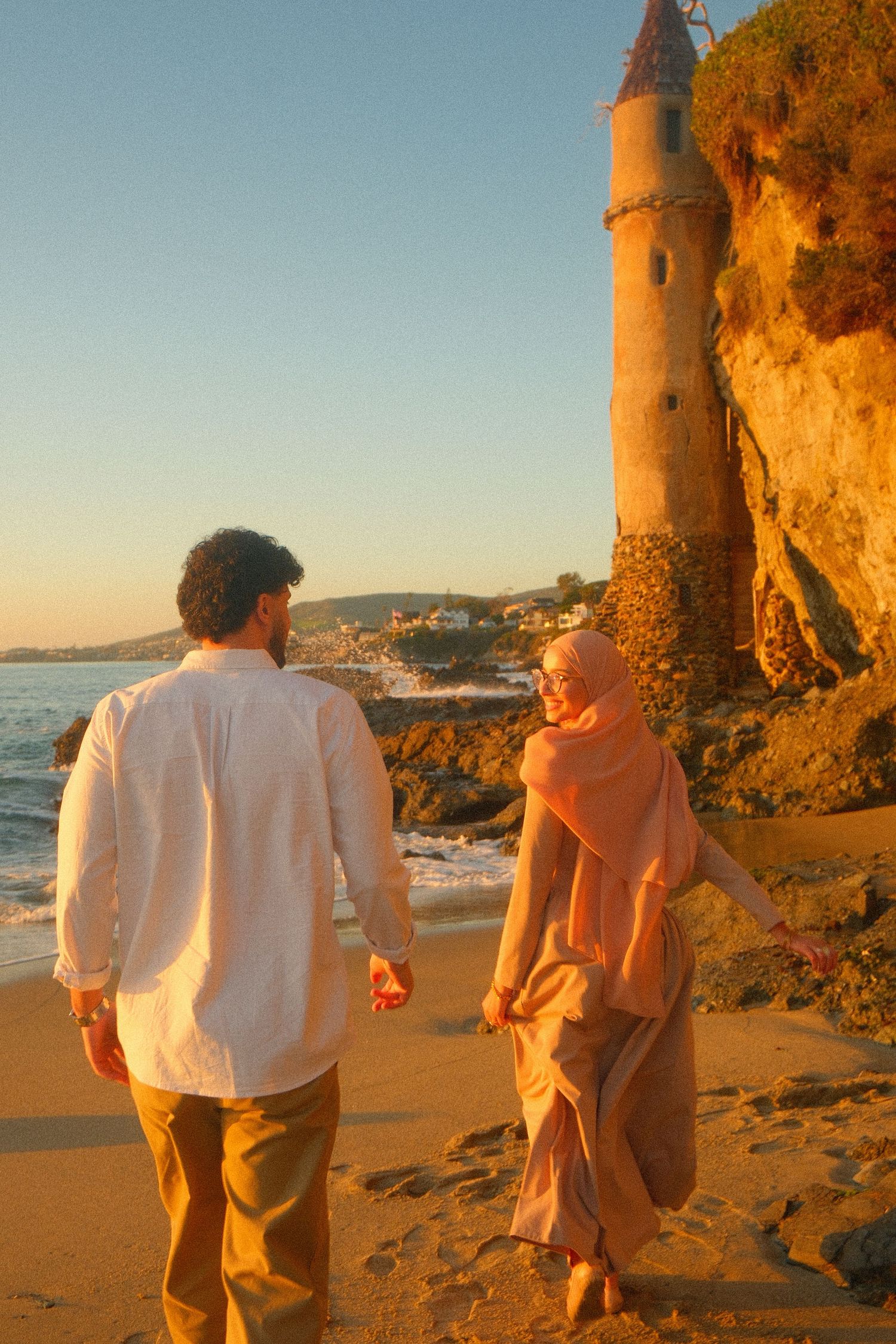 Muslim couple holding hands walking on sandy beach at sunset near rocky cliff.