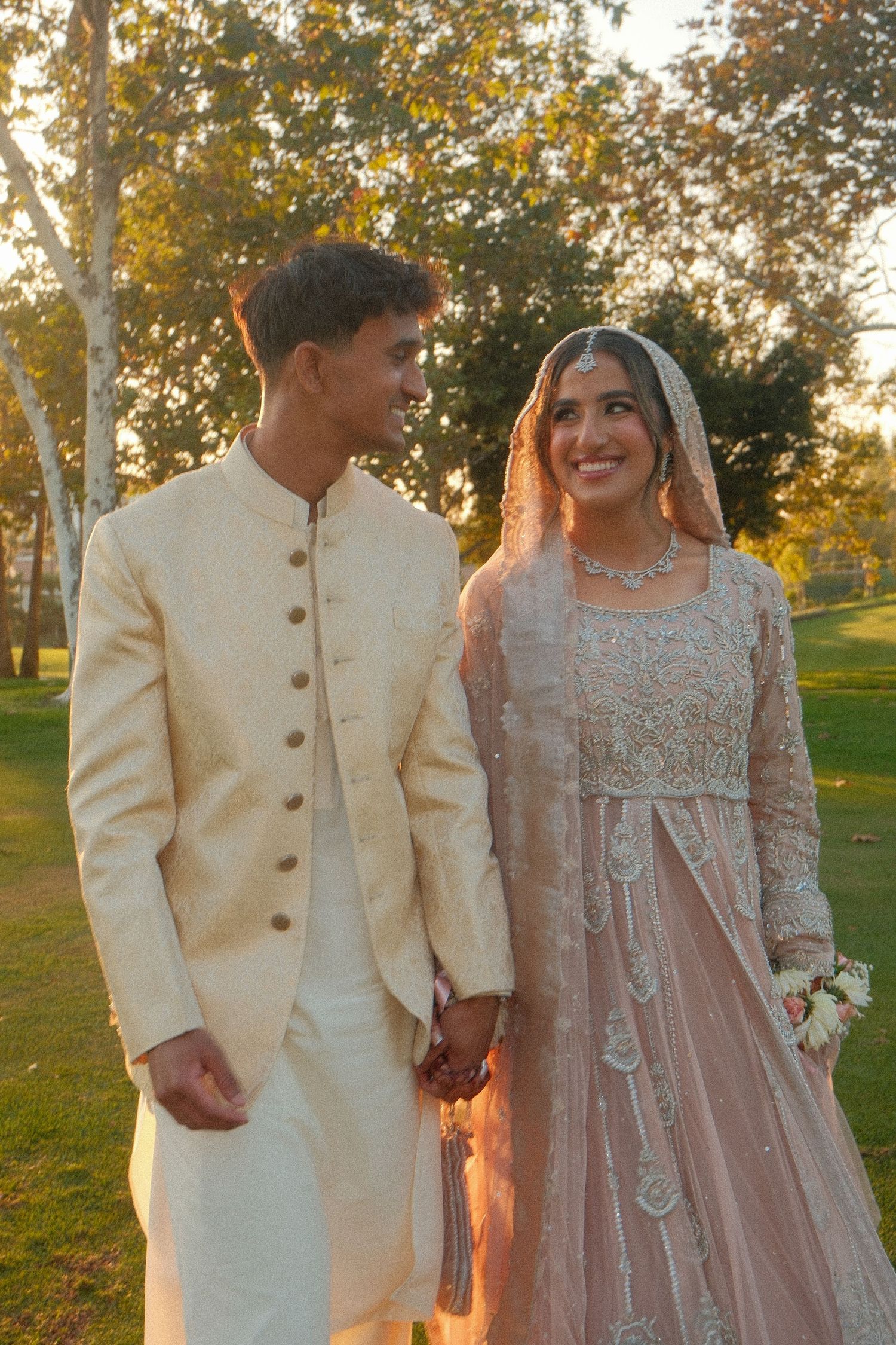 Pakistani bride and groom holding hands outdoors at golden hour, bride in pink embroidered lehenga with dupatta.