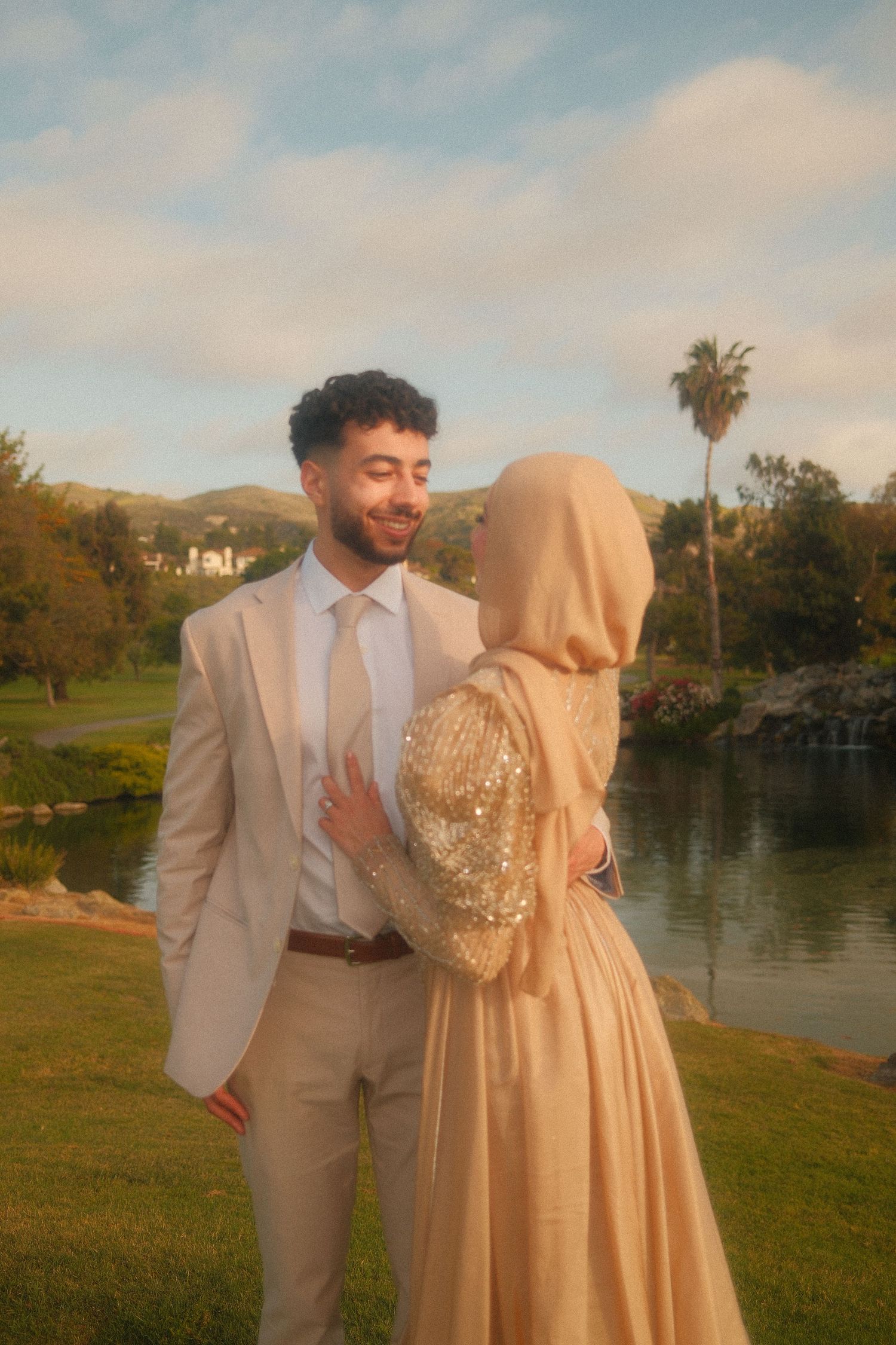 Smiling groom in beige suit gazing at bride in gold sequined hijab dress by a scenic lakeside garden.