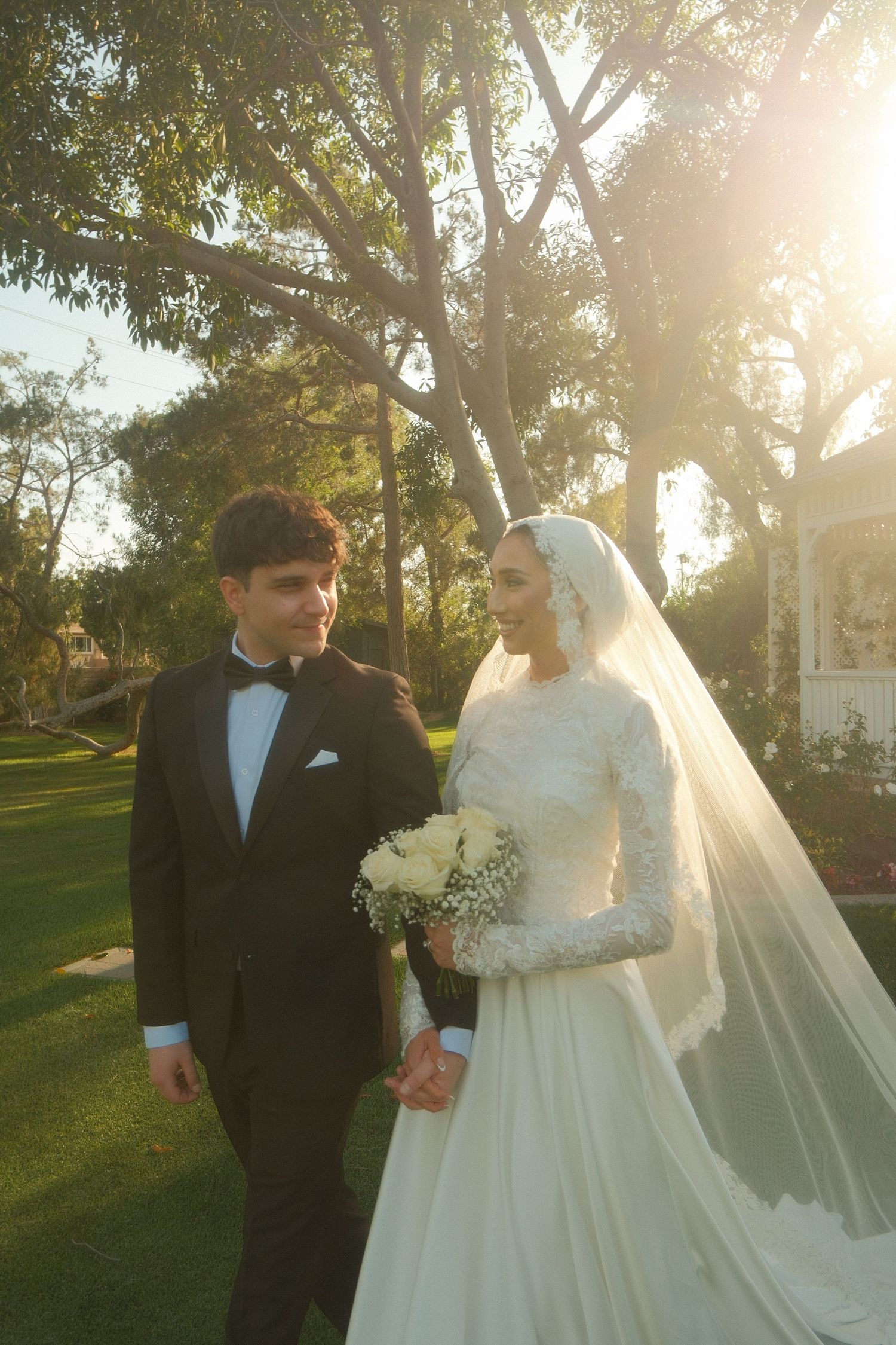 Bride in lace gown with veil holds bouquet beside groom in tuxedo in golden sunset garden.